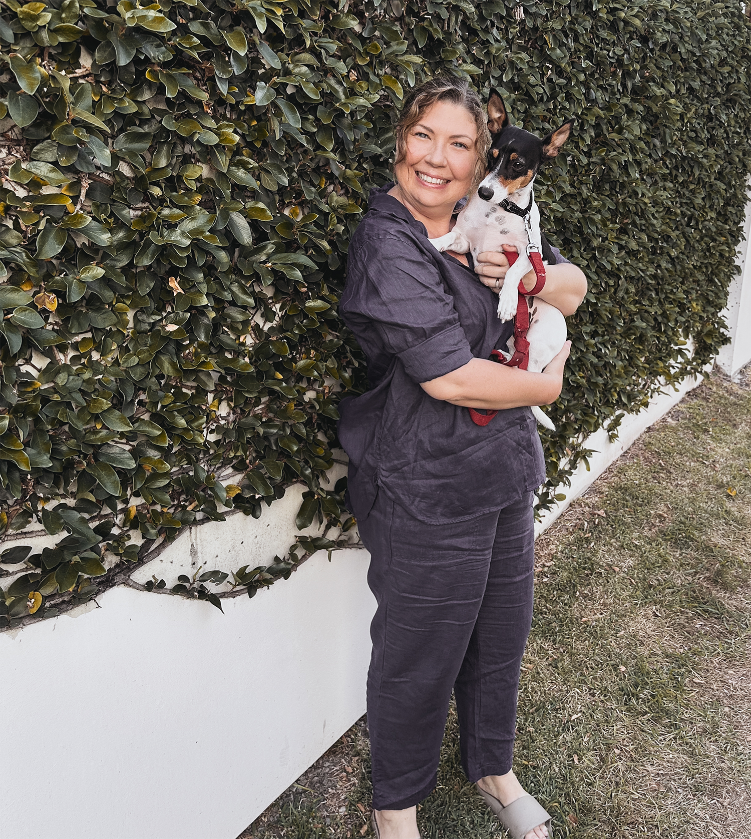 A smiling woman holding a small black and white dog with large ears in front of a lush green hedge, standing on grass outside.