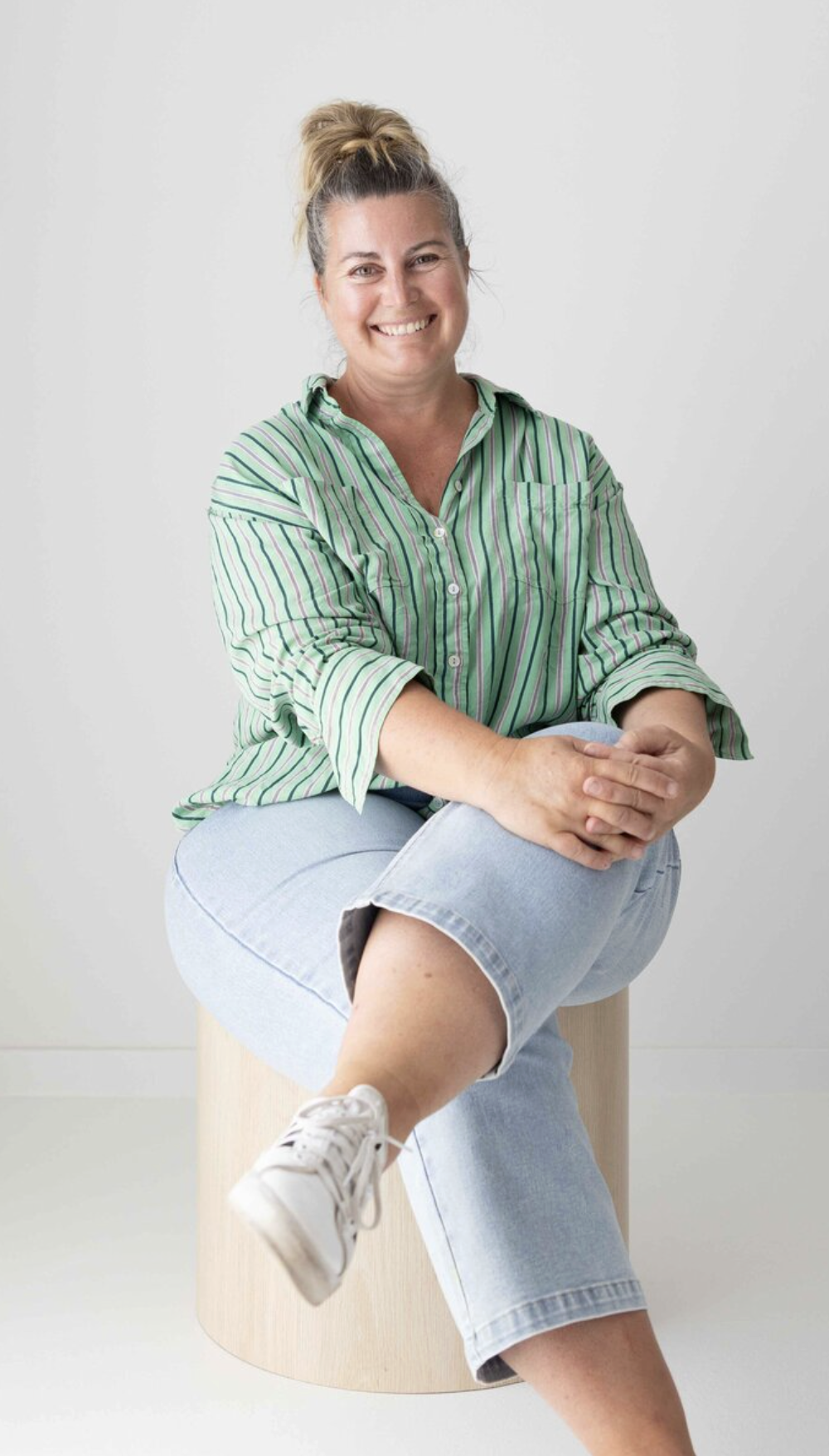 A woman sits cross-legged on a round wooden stool, smiling at the camera. She is wearing a green striped button-up shirt, light blue jeans, and white sneakers. The background is plain white.