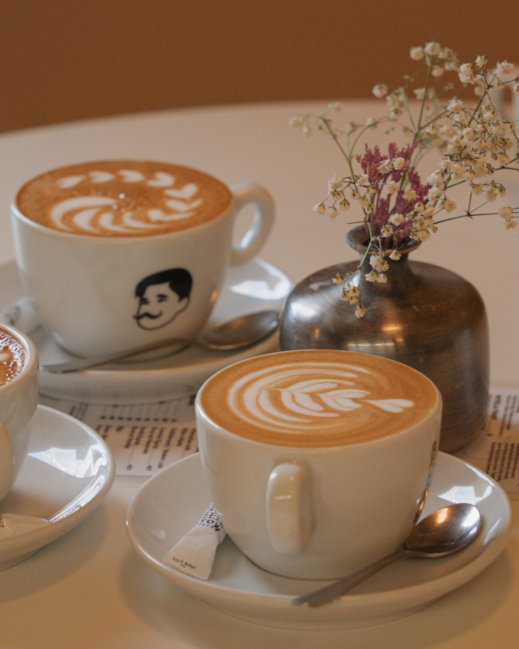 A white cup with a black and white illustration of STOWs log, filled with latte art coffee, on a white saucer. The latte art features a leaf design. The cup is on a tray with silver spoons, a wrapped sugar packet, and a small vase with dried flowers.