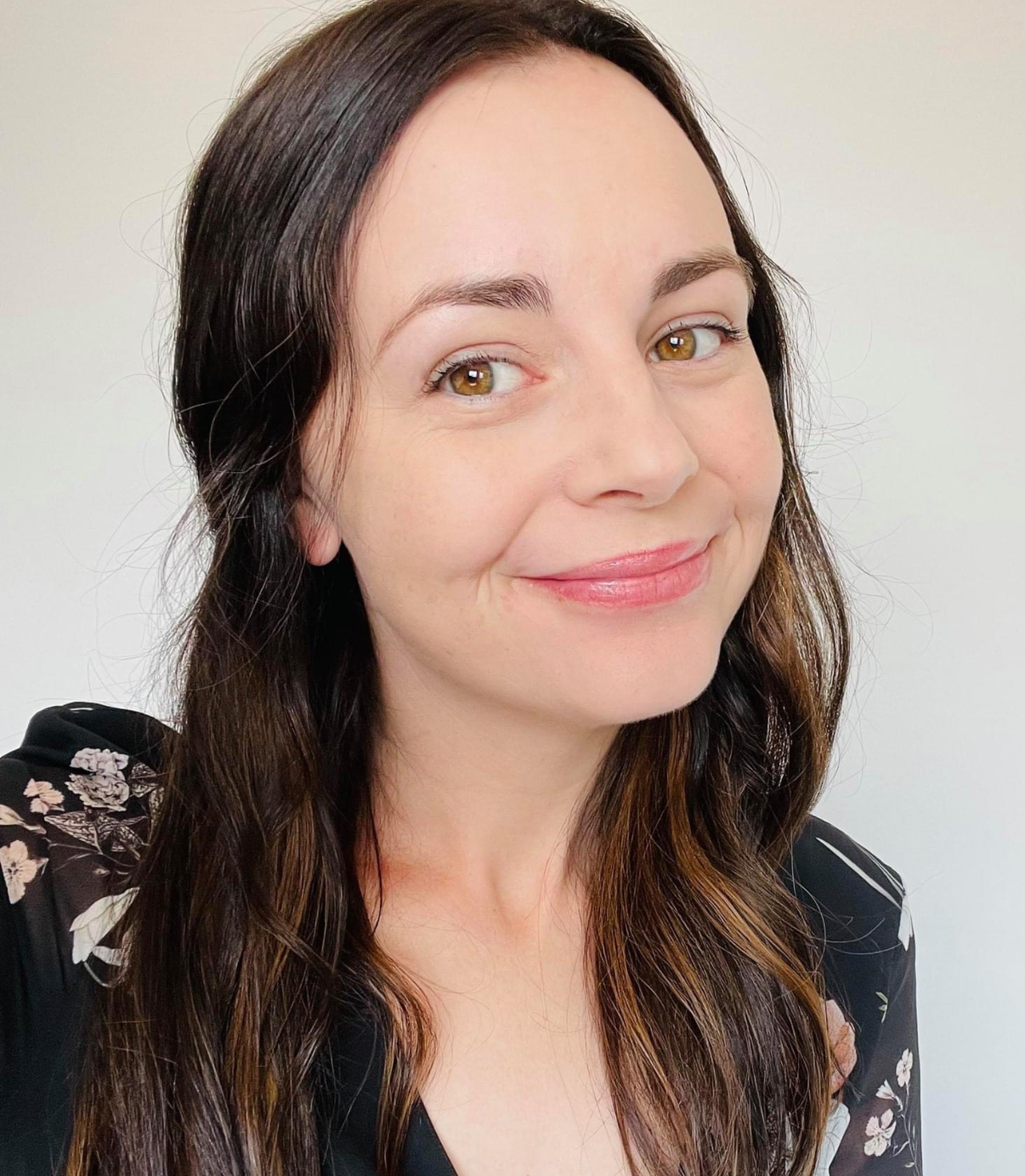 A woman with long, wavy brunette hair and hazel eyes, smiling softly with pink lipstick, wearing a black floral top, standing against a plain light background.