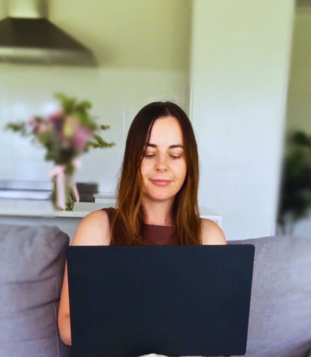 A woman with long brown hair sitting on a sofa, working on a laptop with a blurred flower bouquet in the foreground and a kitchen in the background.