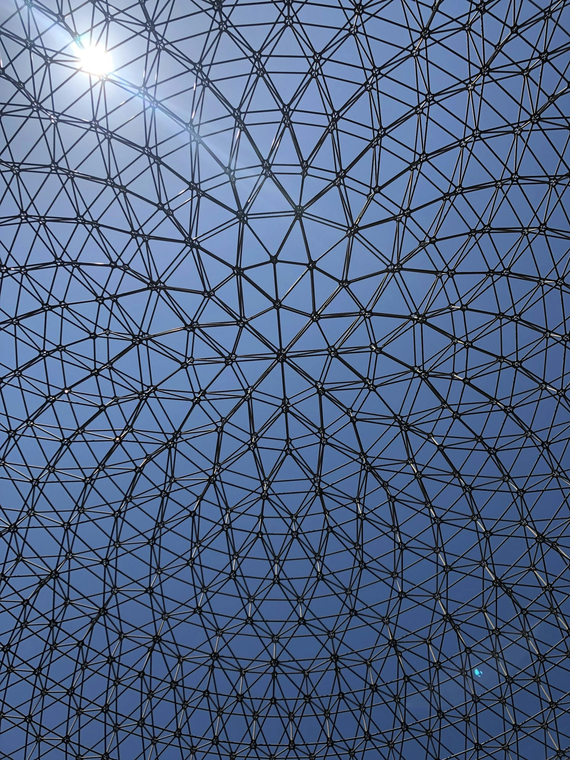 Looking up at a geometric metal dome framework against a bright blue sky with the sun shining through.