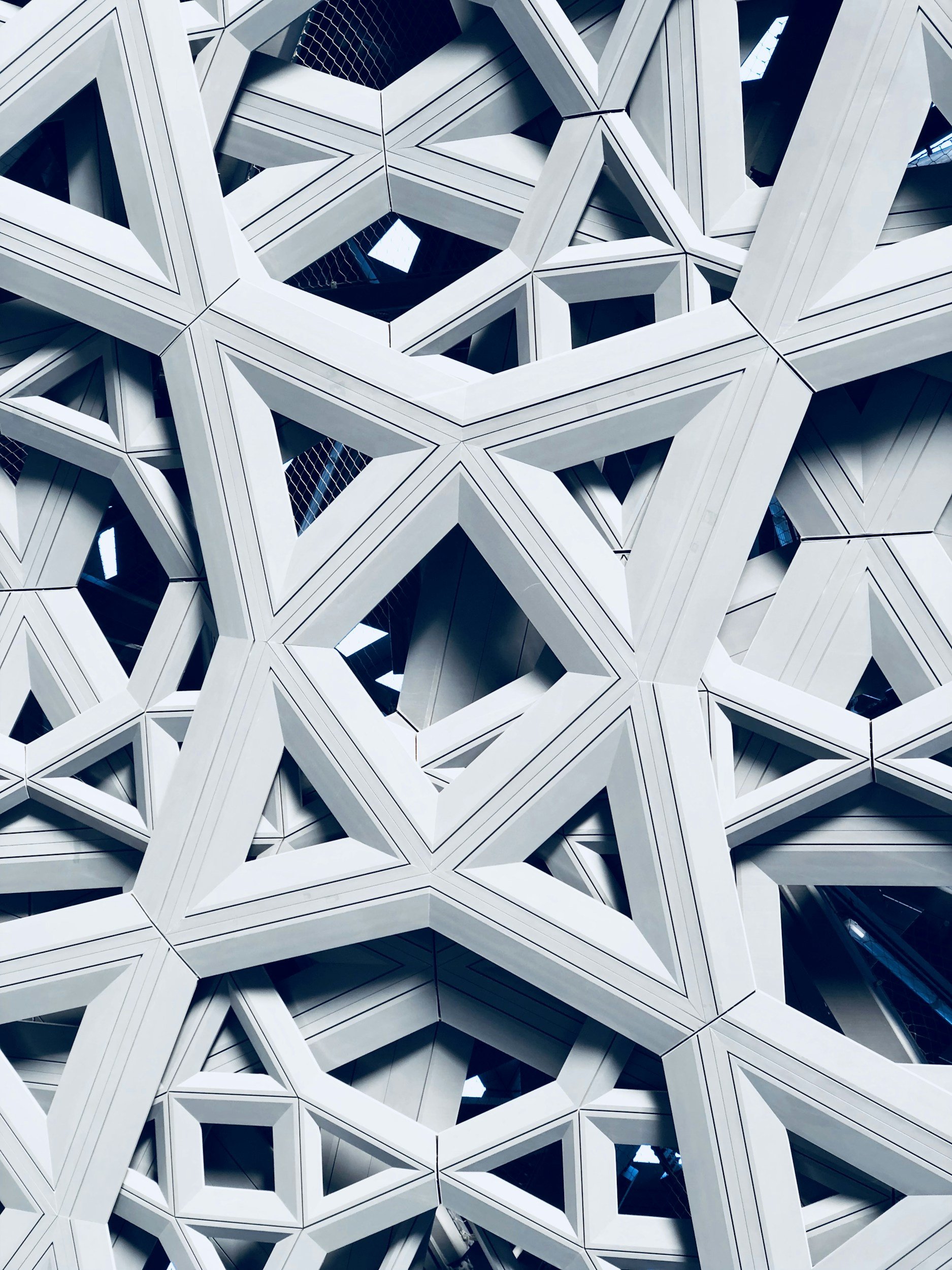 A detailed view of a modern architectural facade made of interconnected geometric shapes and patterns, primarily white, with some dark and reflective surfaces visible within the structures.