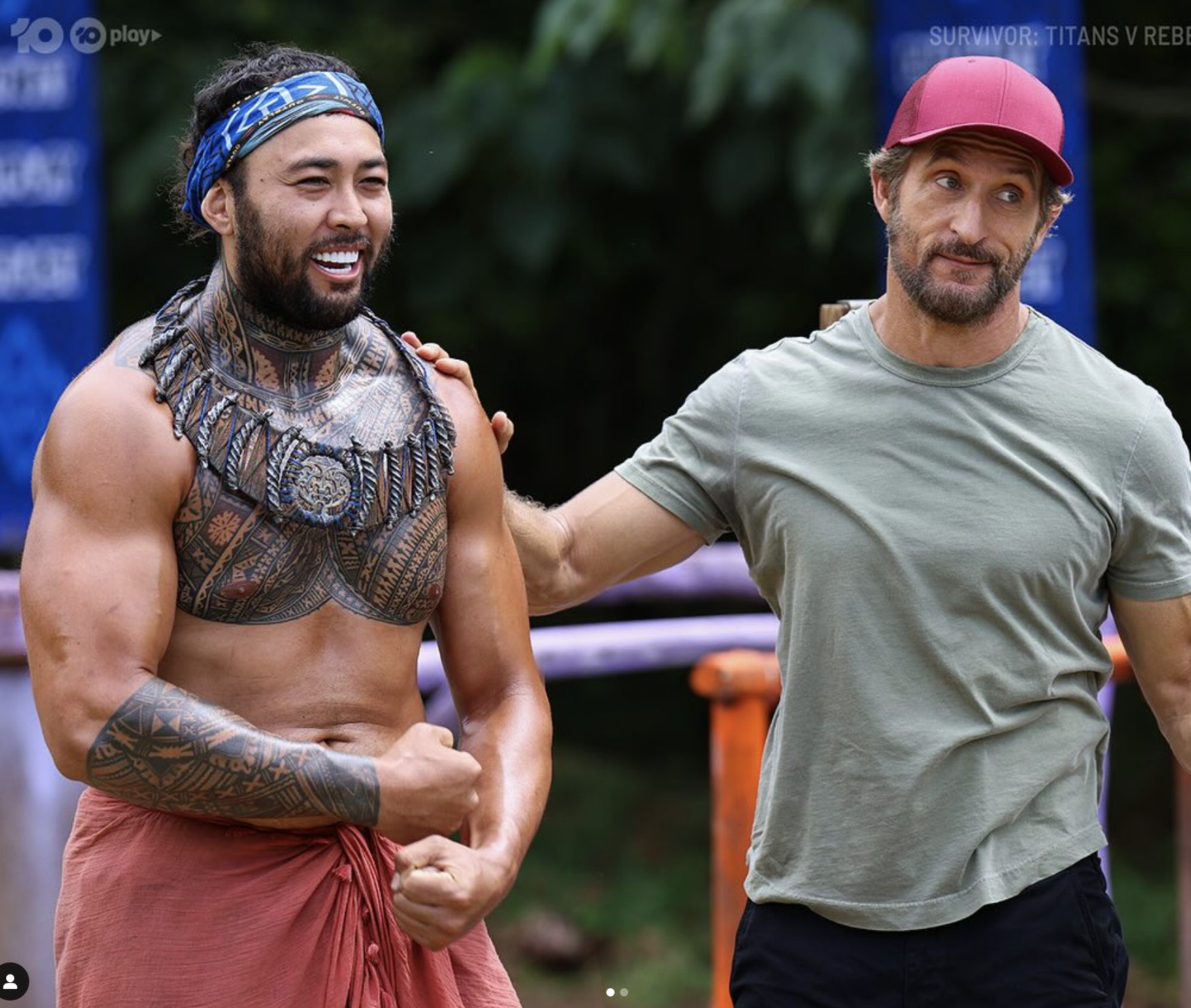 Two men outdoors, one with tribal tattoos and traditional attire, the other in a casual t-shirt and cap, standing together.