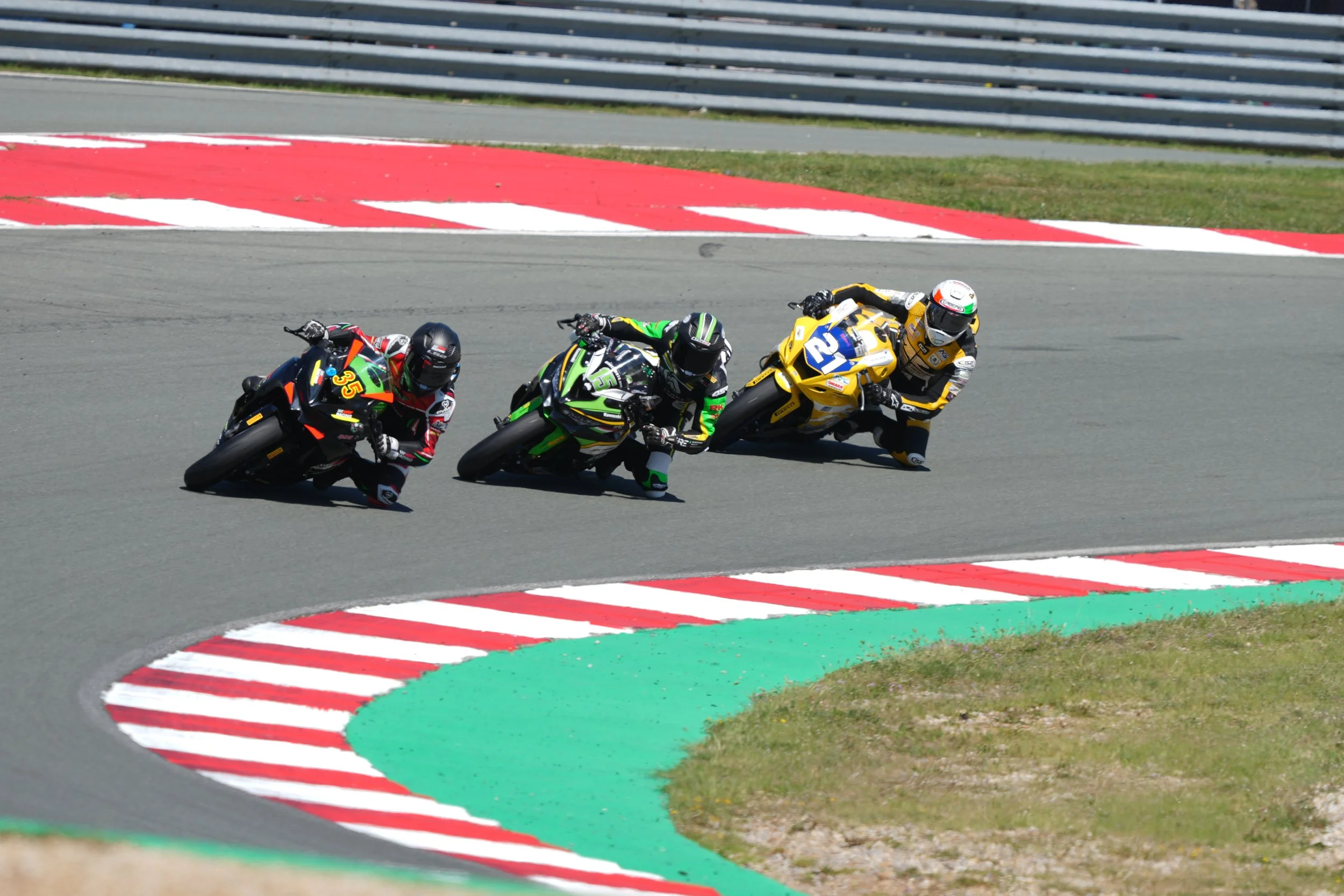 Three motorcyclists racing around a track, leaning into a turn with red and white striped curbing on the edge of the asphalt.