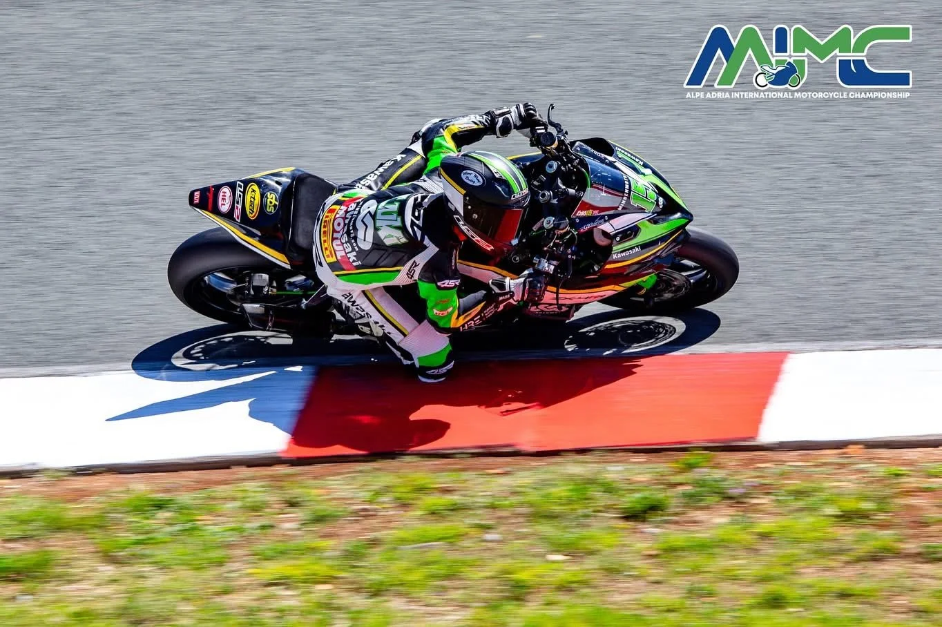 A motorcycle racer in green and black gear leaning into a corner on a racing track at the Alpe Adria International Motorcycle Championship, with the logo in the top right corner.