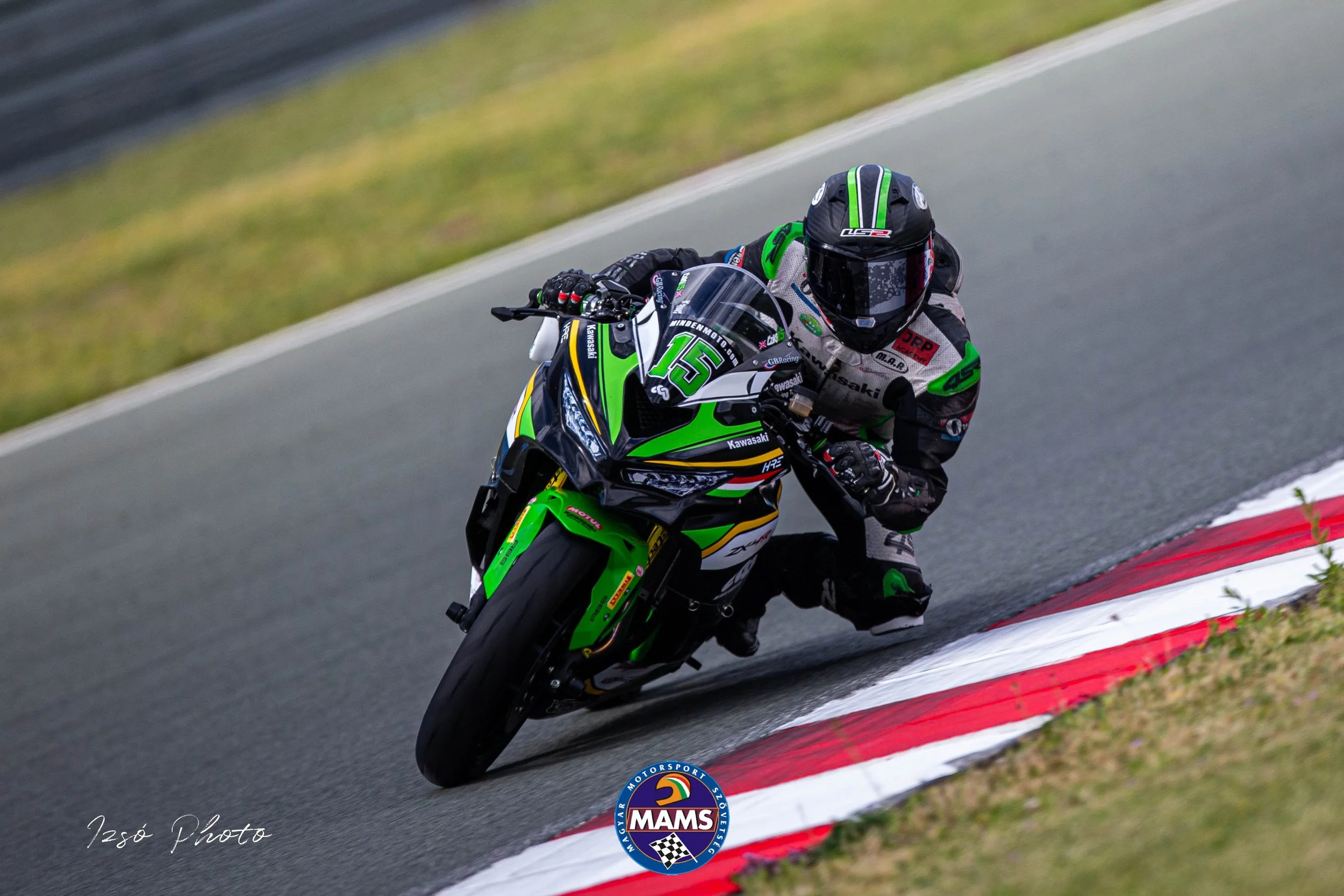Motorcycle racer in racing gear leaning into a turn on a racetrack, with green and black motorcycle and '15' on the front, logo of MAMS at the bottom, and green grass on side of track.