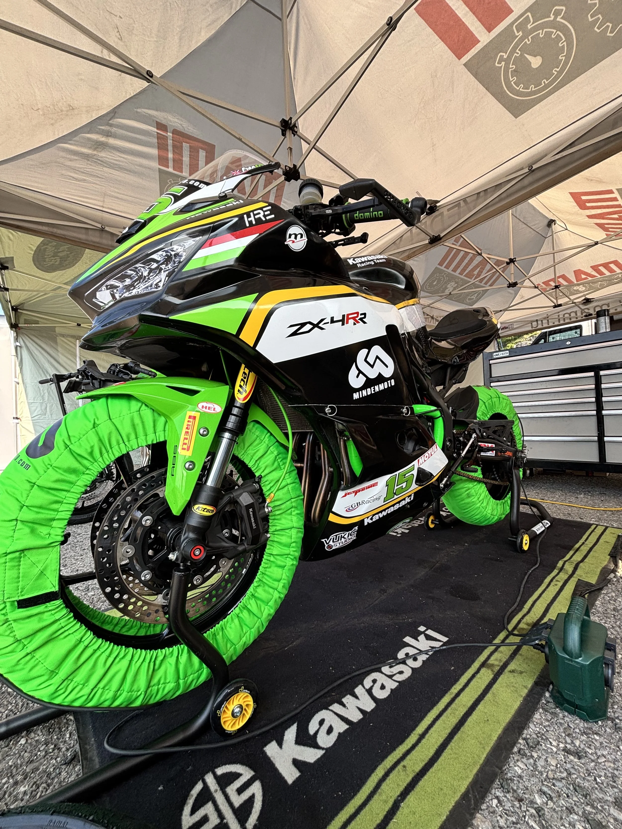 Kawasaki racing motorcycle with green tire warmers inside a tent at a race track.