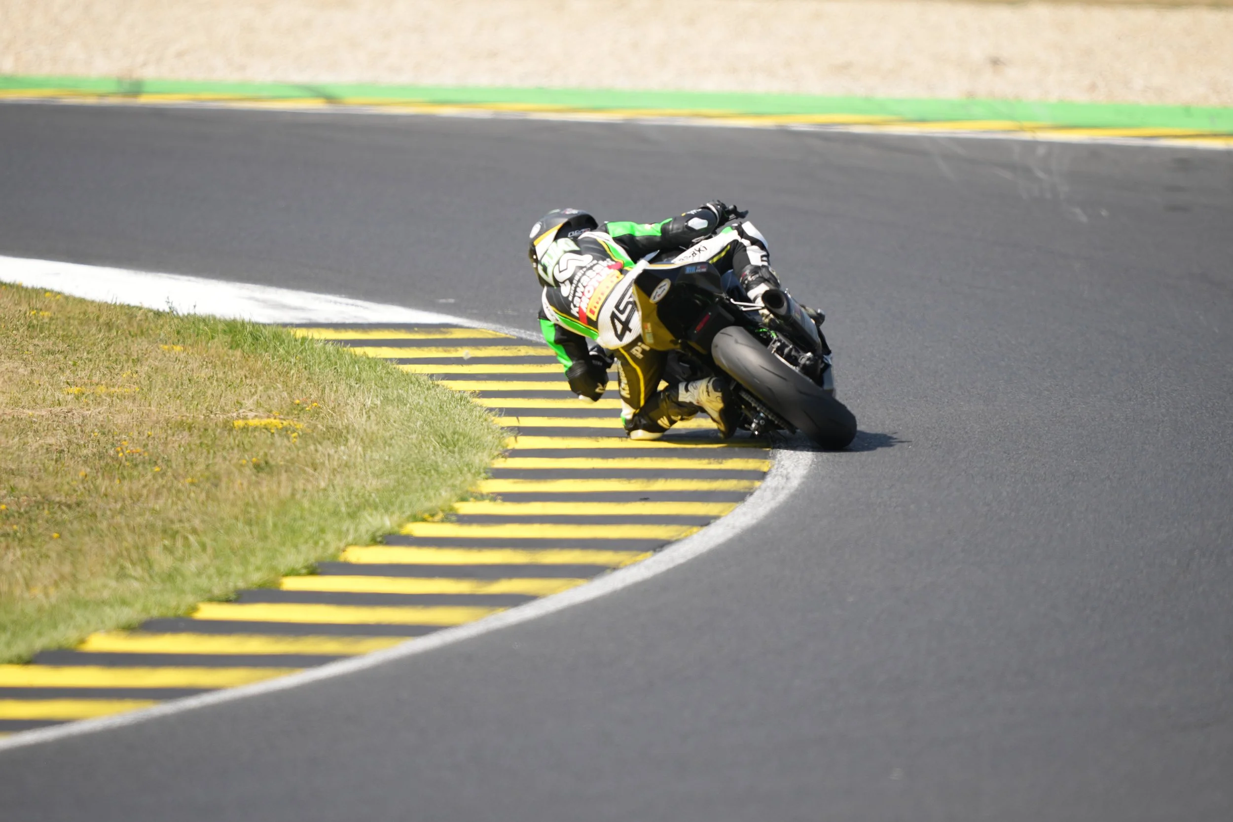 Motorcycle rider leaning into a turn on a race track, with yellow and black striped curb and grassy area nearby.