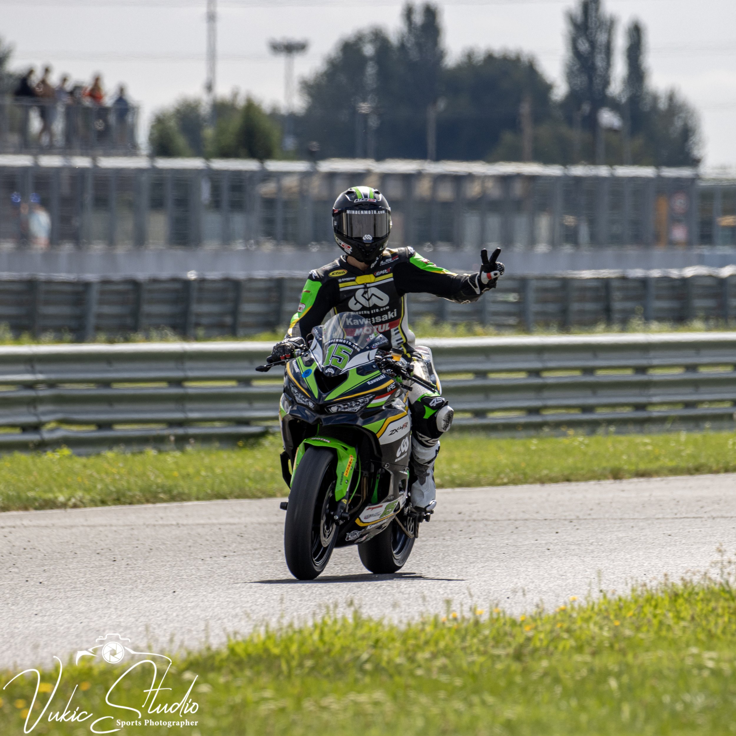 Motorcycle racer on a green and black Kawasaki motorcycle making a peace sign while riding on a race track.