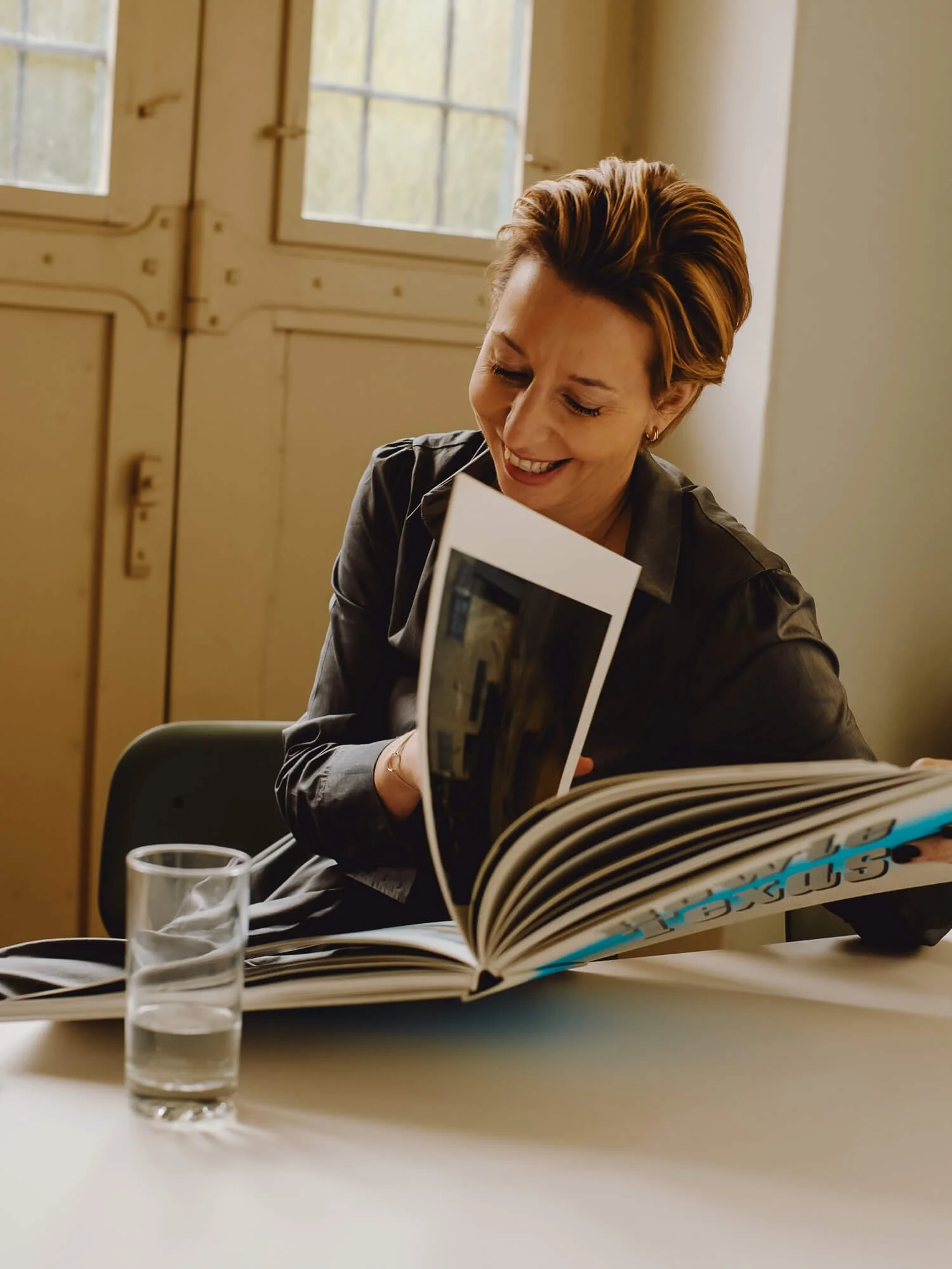 Woman with reddish-brown hair wearing a dark blazer, smiling while looking at an open magazine or portfolio at a white table. A glass of water sits beside her, with wooden doors and windows visible in the background.