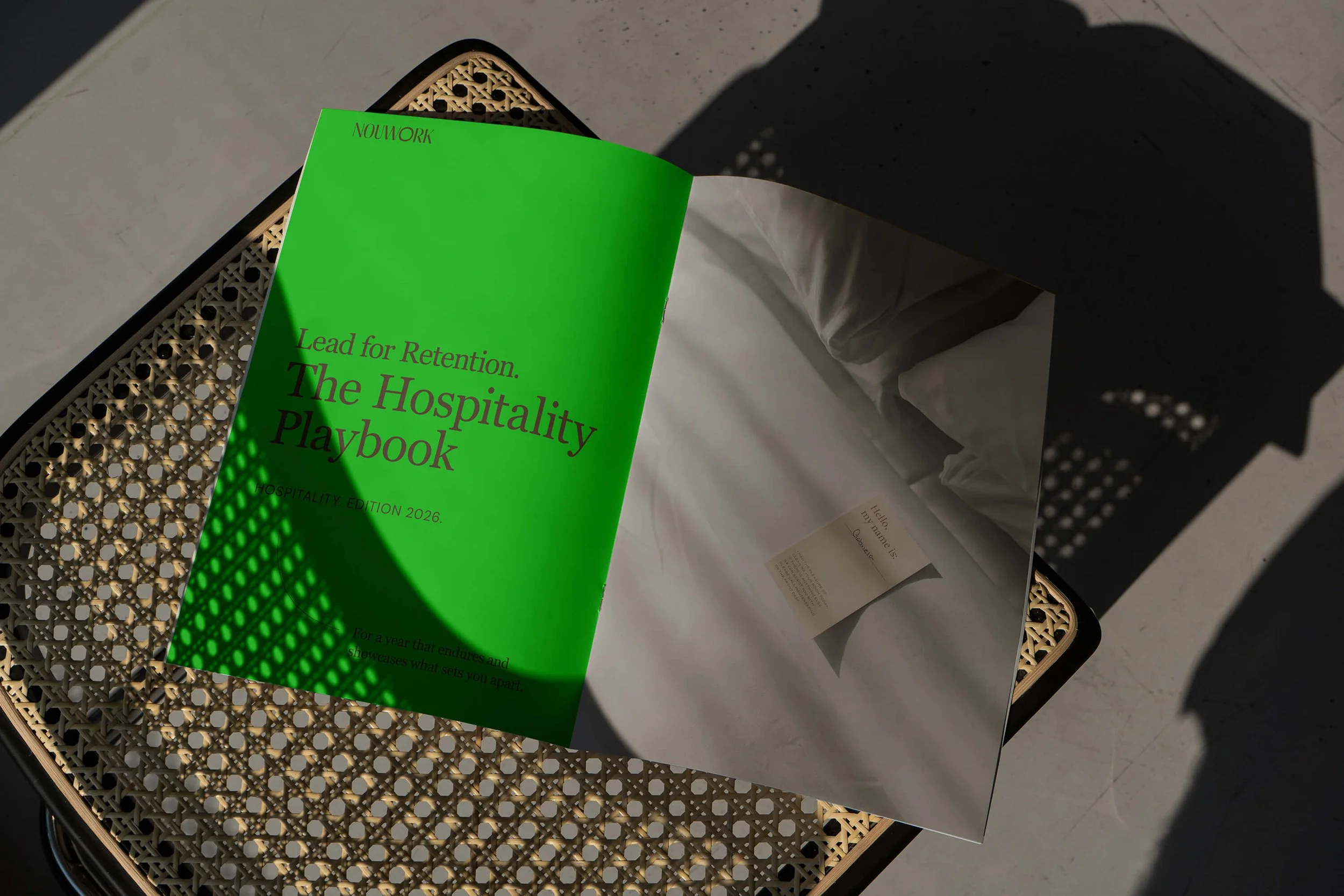 A green booklet titled 'The Hospitality Playbook' in German and English, placed on a wooven brown chair showing the booklet, against a gray background with shadows