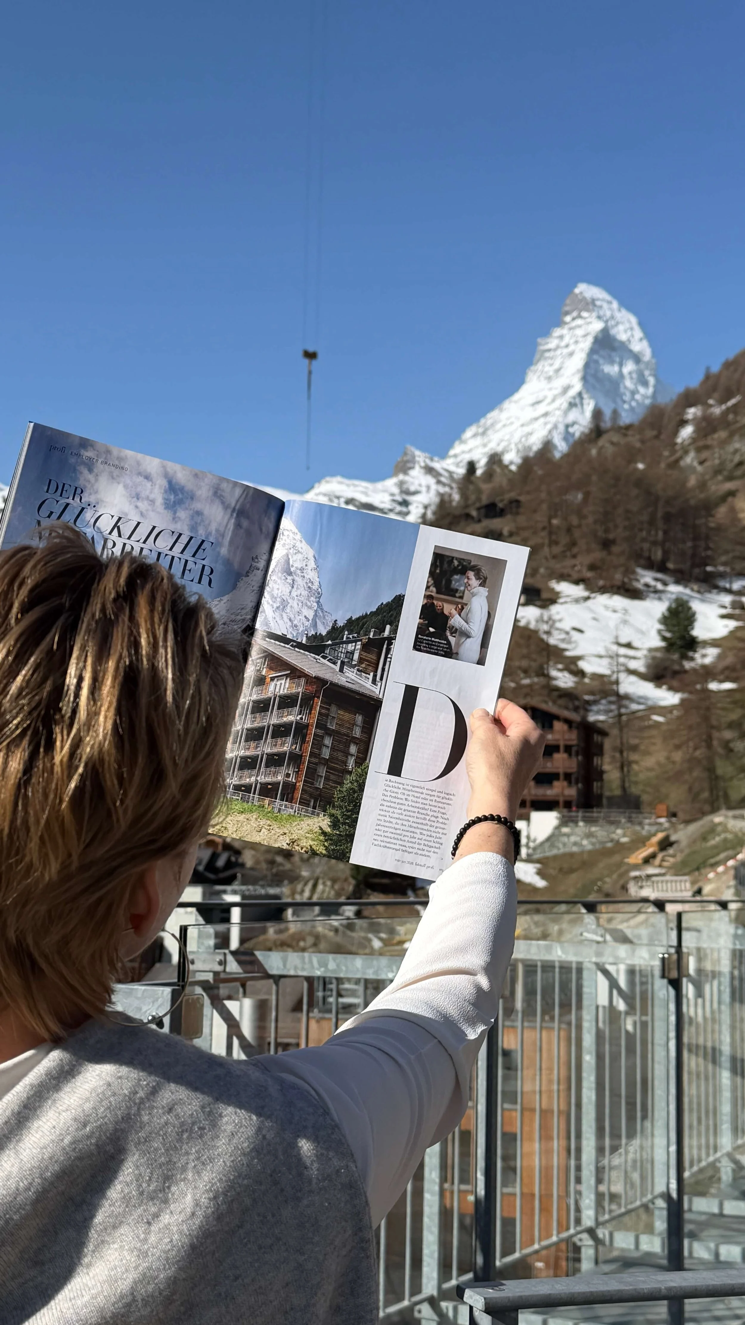 Person holding open a magazine featuring an article titled 'Der Glückliche Mitarbeiter' (The Happy Employee), with the iconic Matterhorn mountain in the background under a clear blue sky, Zermatt, Switzerland.