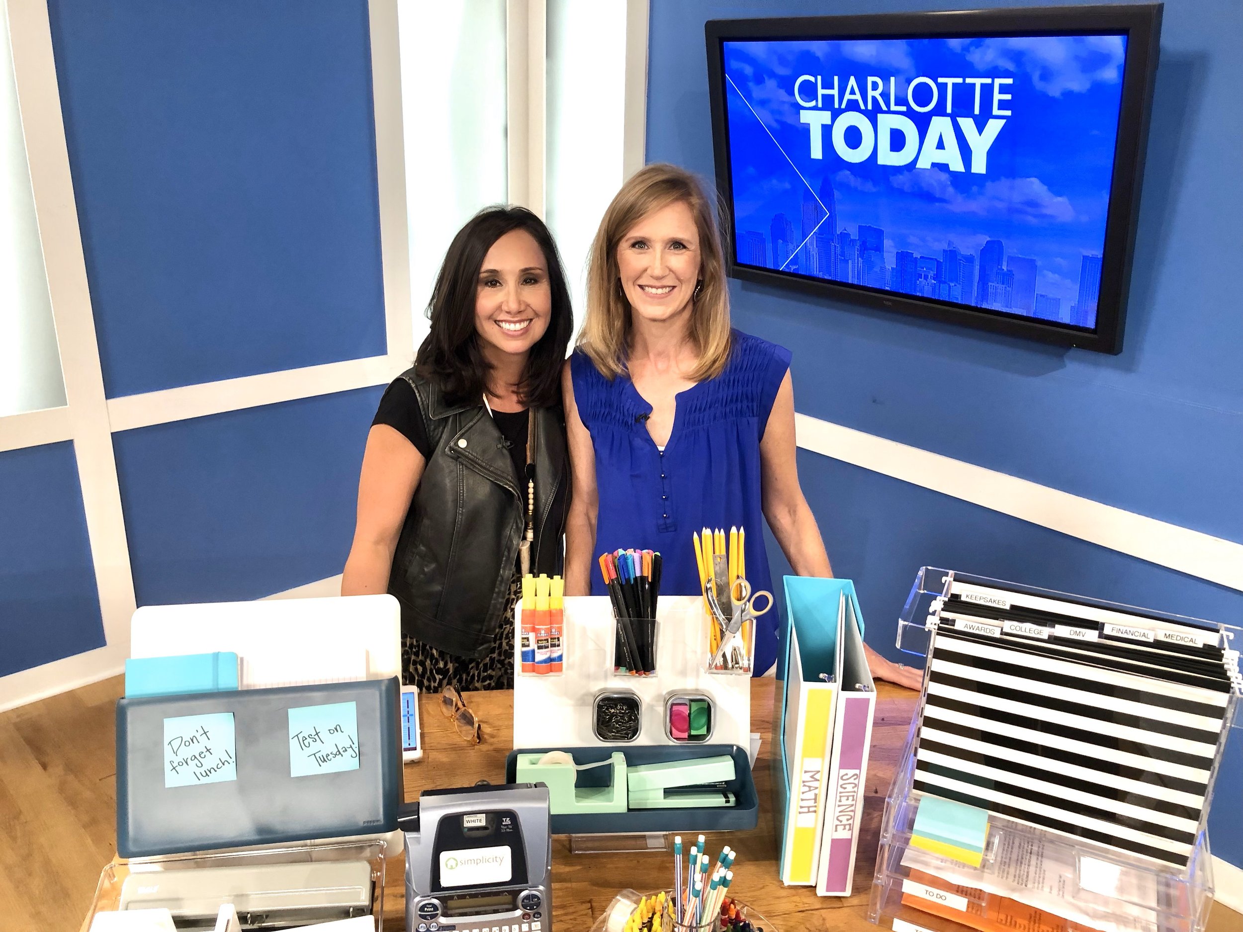 Two women standing behind a desk with organized office supplies, including binders, pens, sticky notes, and a label maker. The backdrop displays a screen with the text "Charlotte Today."
