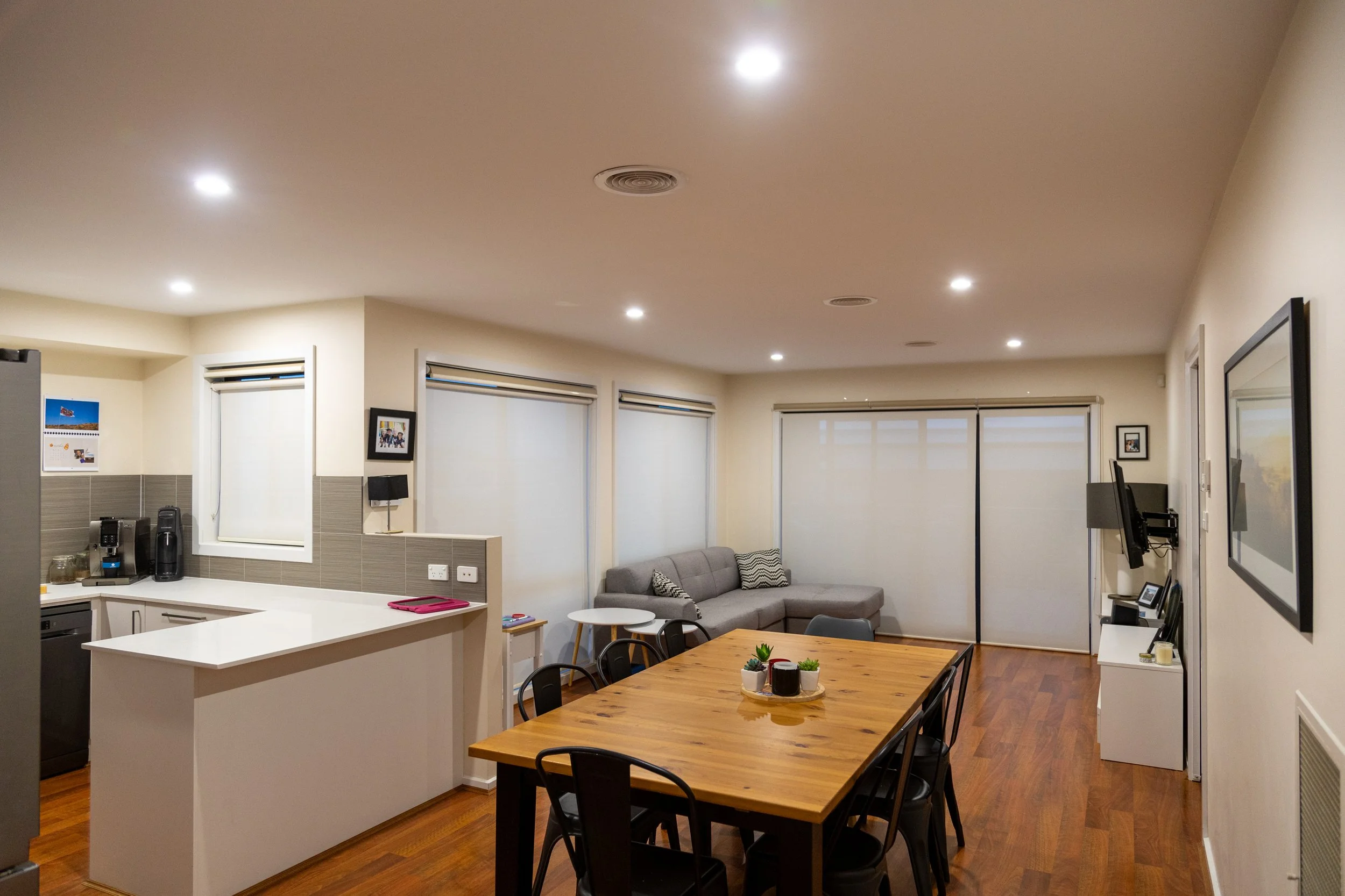 Modern living room and kitchen with wooden floors, a dining table, chairs, a sectional sofa, and a small kitchen area with a countertop and appliances.