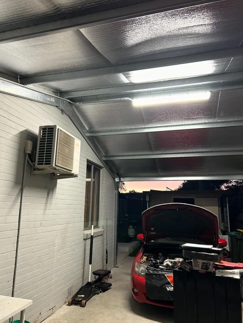 Garage interior with a red car under maintenance, hood open, tools visible, air conditioning unit on the wall, and metal roofing.