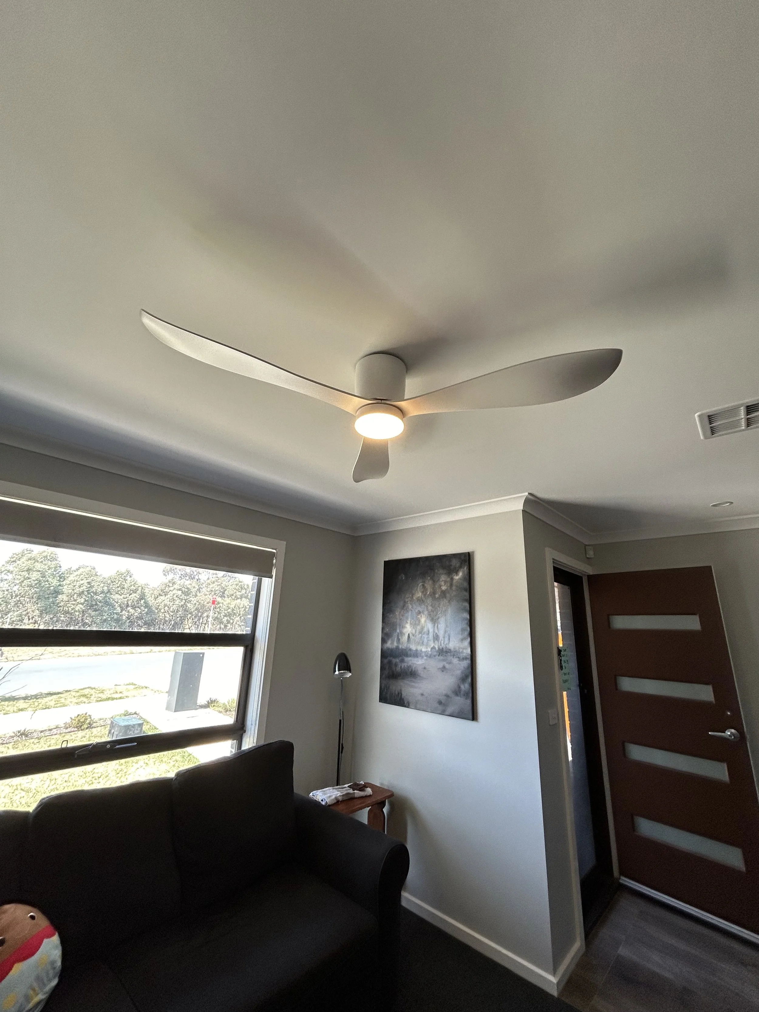 Interior of a room with a ceiling fan, a window with a view, a couch, a floor lamp, a wall painting, and a door.