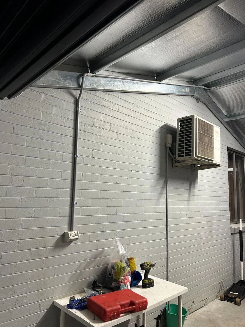 Indoor workspace with wall-mounted air conditioning unit, brick wall, table with tools, and metal roof beams.