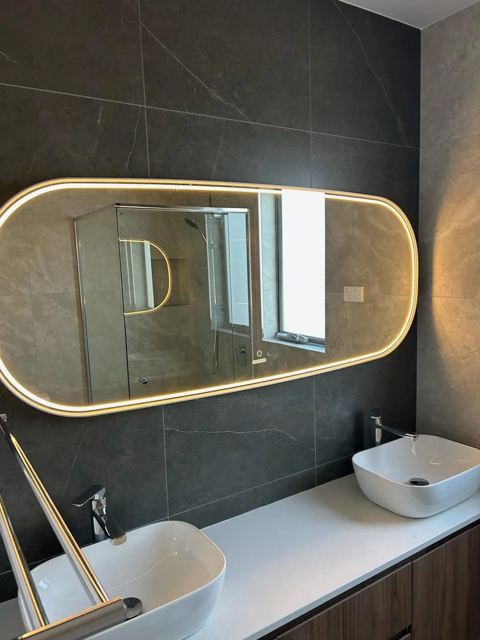 Modern bathroom with oval backlit mirror, dual sinks, dark tile wall, and glass shower enclosure.