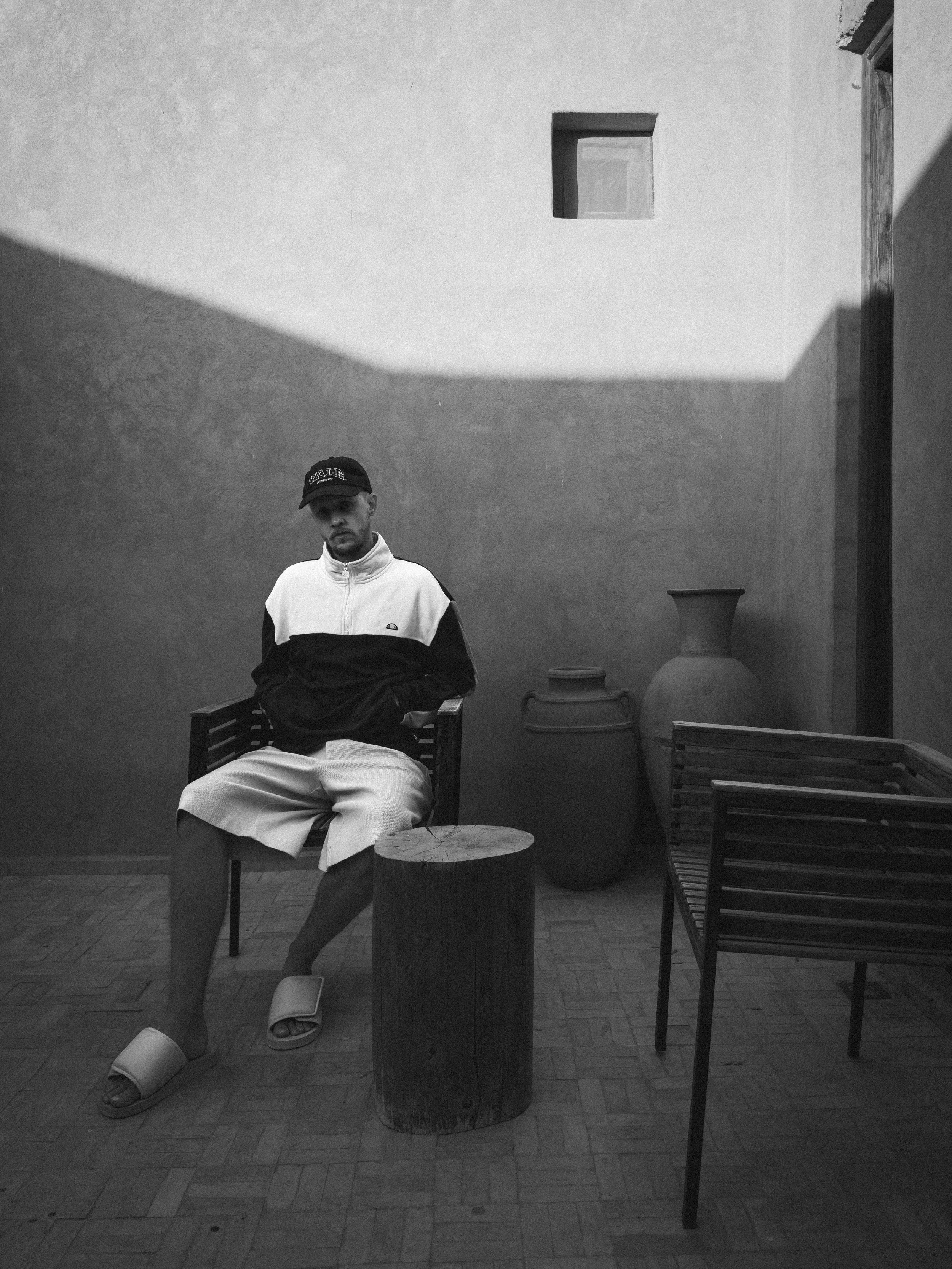 A man sitting on a chair outdoors with large vases nearby, wearing a cap, jacket, shorts, and slippers, in a shaded area of a courtyard or patio with a textured wall and a small window.