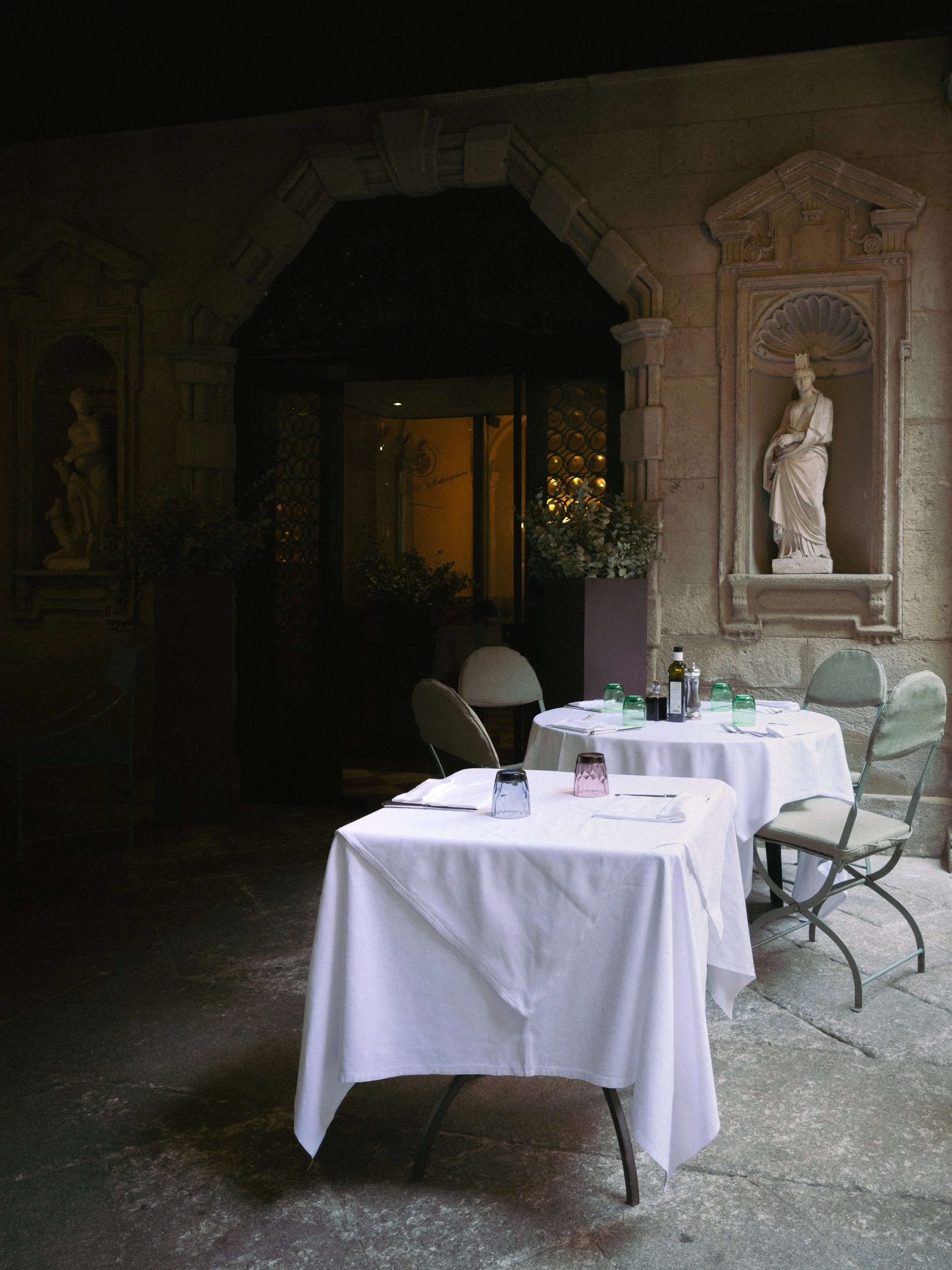 Two round tables with white tablecloths and chairs in a restaurant with stone walls and statues