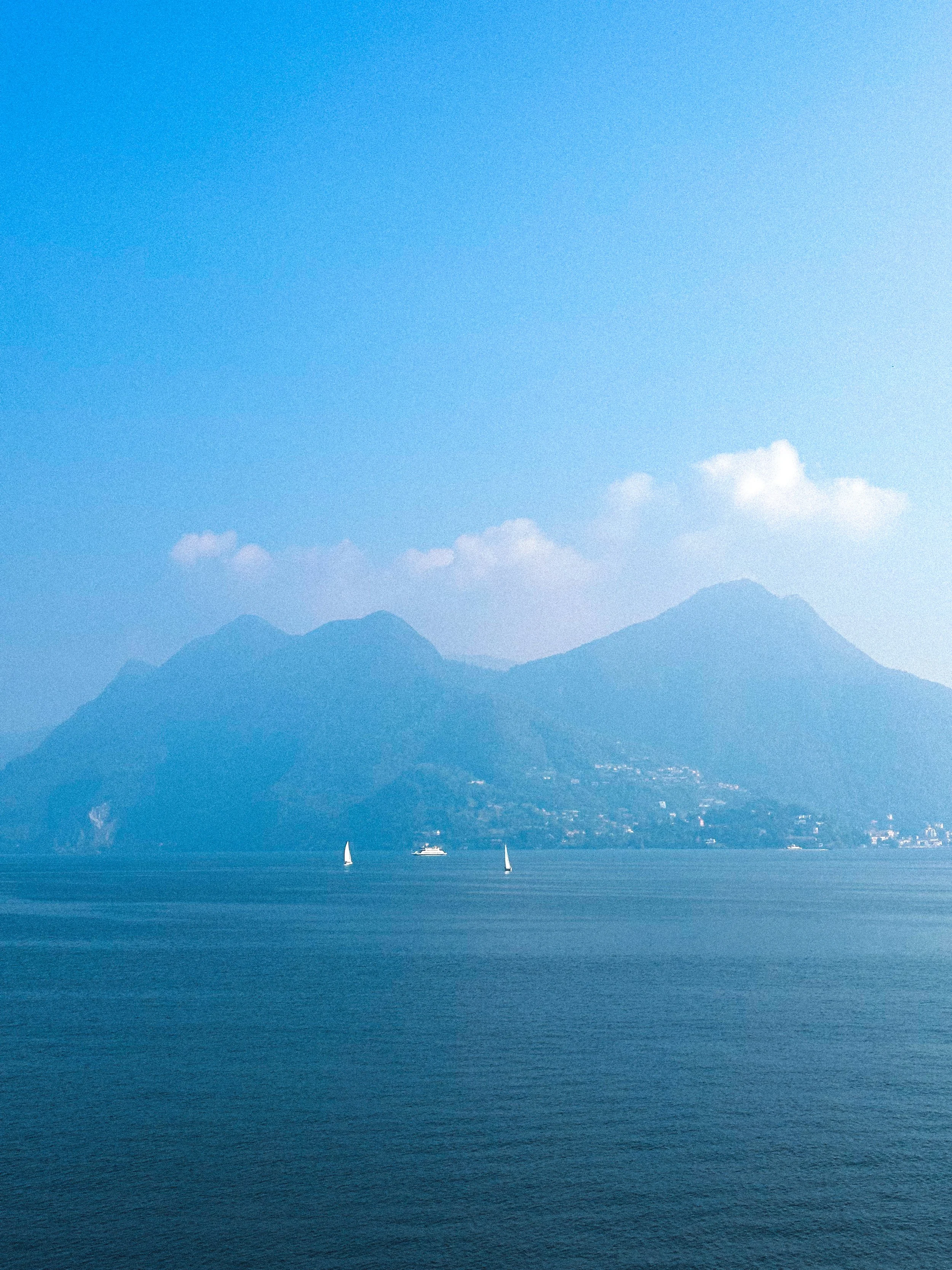 Scenic view of a large lake with sailboats, mountains in the background, and a blue sky with a few clouds.