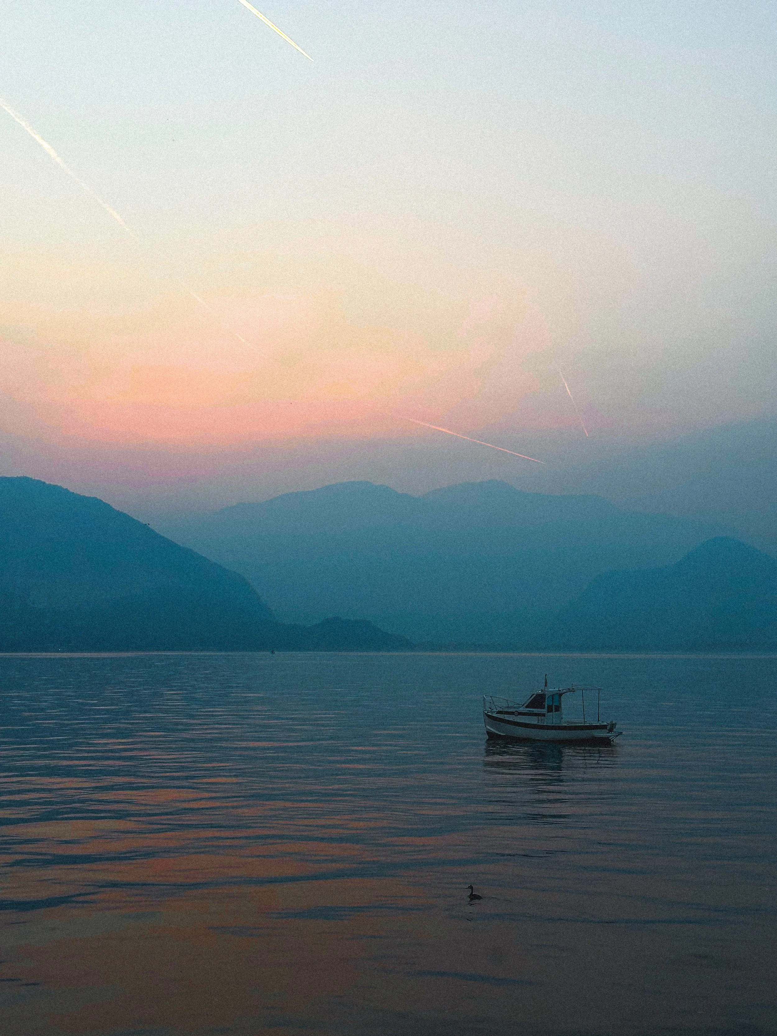 A peaceful lake scene at sunset with a small boat floating on calm water, mountain ranges in the background, and a colorful sky with pink and blue hues.
