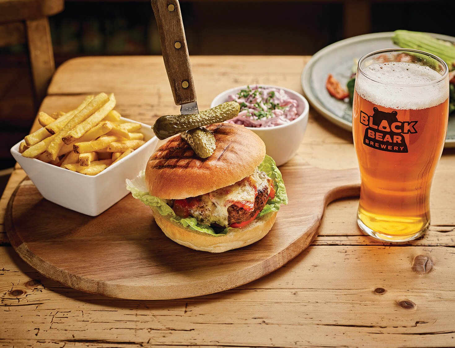 An image of a pint of Black Bear Brewery Ale together with a cheeseburger and fries on a wooden serving platter