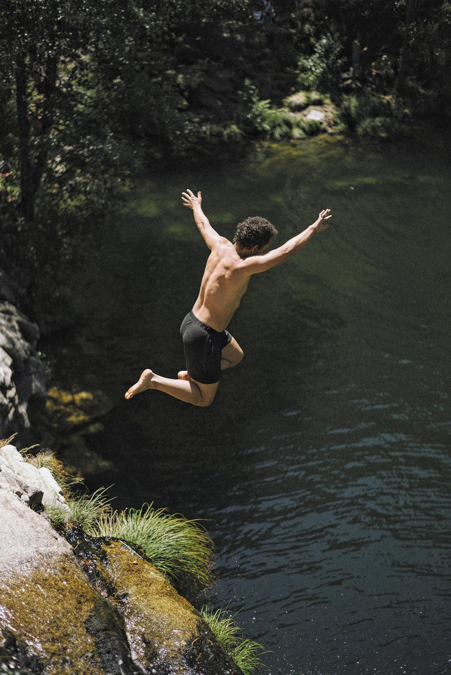 Pedro jumping shirtless into mountain lake from granite waterfall stone, arms spread wide in embodied freedom