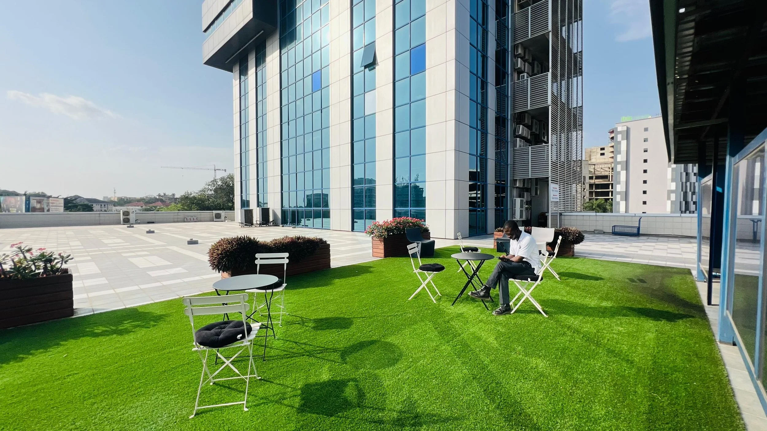 Man sitting on chair on rooftop patio with artificial grass, surrounded by tables, chairs, and potted plants, with modern glass building in background.