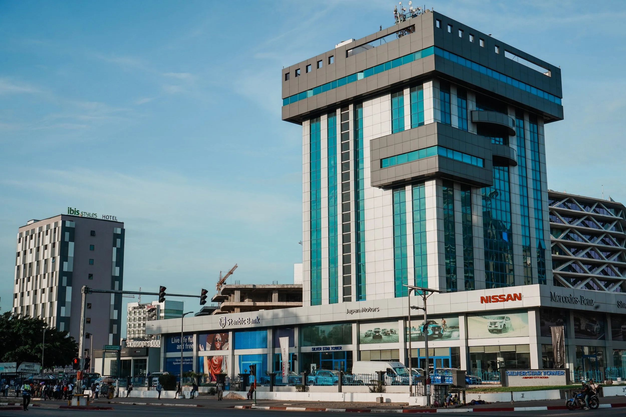 Cityscape with tall modern buildings, including a high-rise with blue glass windows and a unique architectural design, alongside an Ibis Styles Hotel and a Mercedes-Benz showroom. Cars and pedestrians are visible on the street.