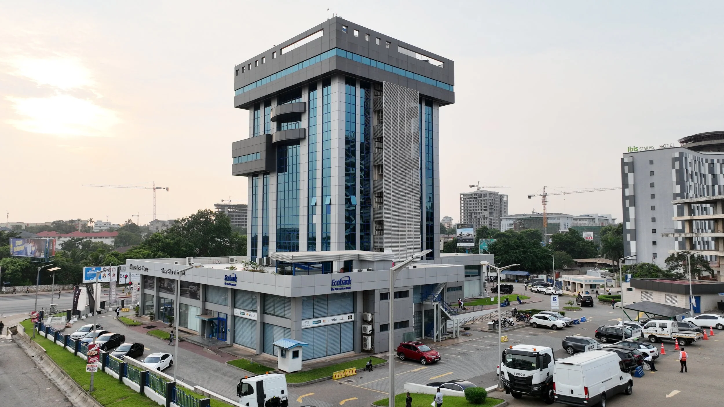 Tall modern office building with reflective glass windows, a shopping center on the ground floor, and a parking lot with cars in the foreground, in an urban area during daytime.