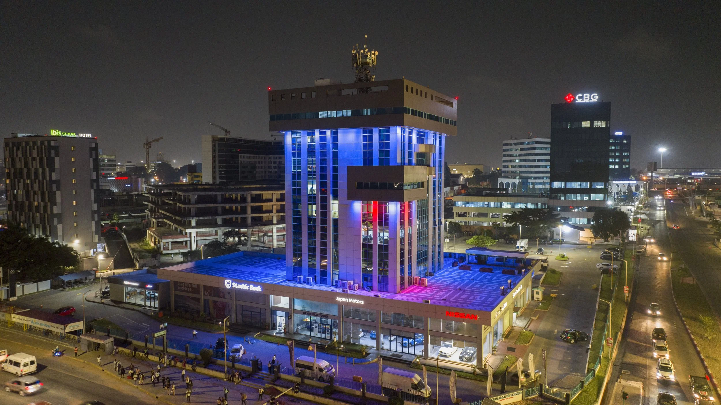 Nighttime cityscape featuring a tall building with blue and purple lighting atop a car dealership, with surrounding buildings and a busy street with cars and pedestrians.
