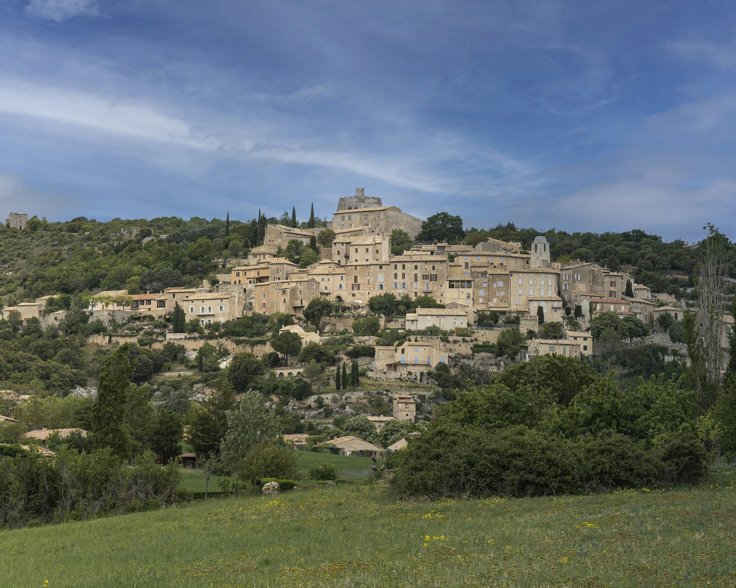 A hillside village with old stone buildings, surrounded by greenery and trees under a partly cloudy sky.