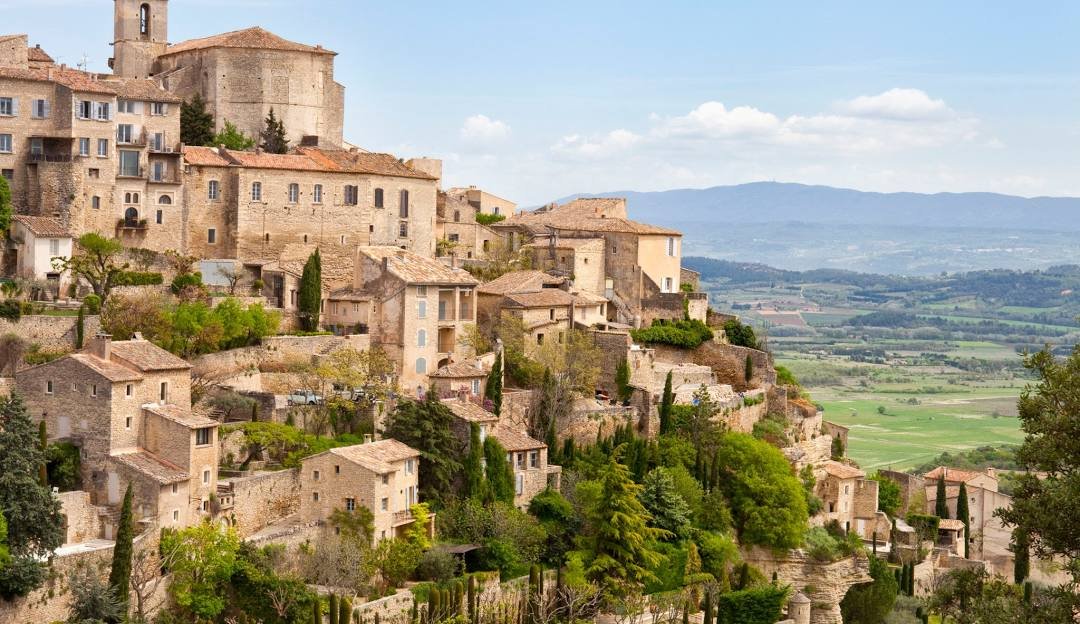 A hillside village with stone buildings and terracotta roofs, surrounded by greenery and trees, with a rural landscape and mountains in the background under a partly cloudy sky.
