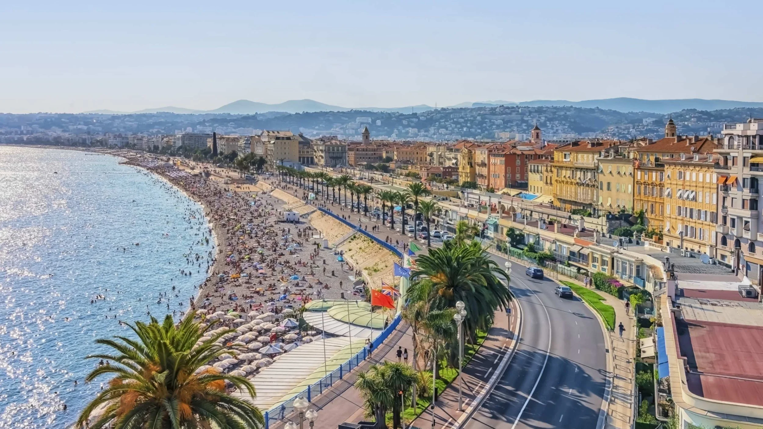 Beach with crowded shoreline and umbrellas, palm trees, seaside promenade with flags, and colorful buildings along a coastal road with cars.