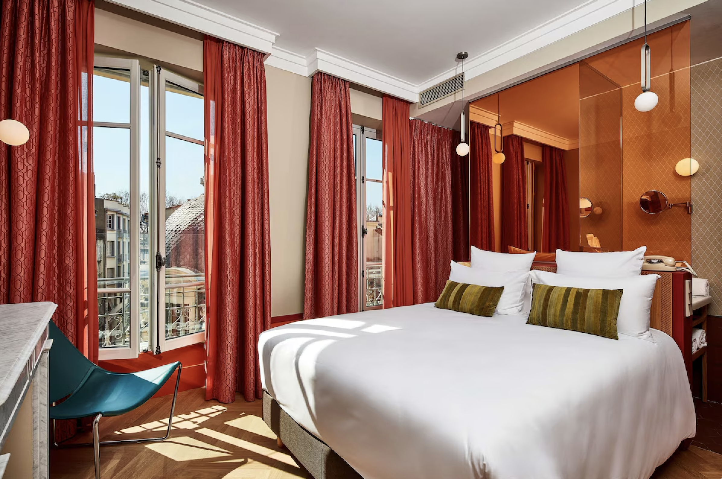 Hotel room with large bed, white linens, and two green pillows. Red curtains and orange wall accents. Large windows with sunlight and city view.