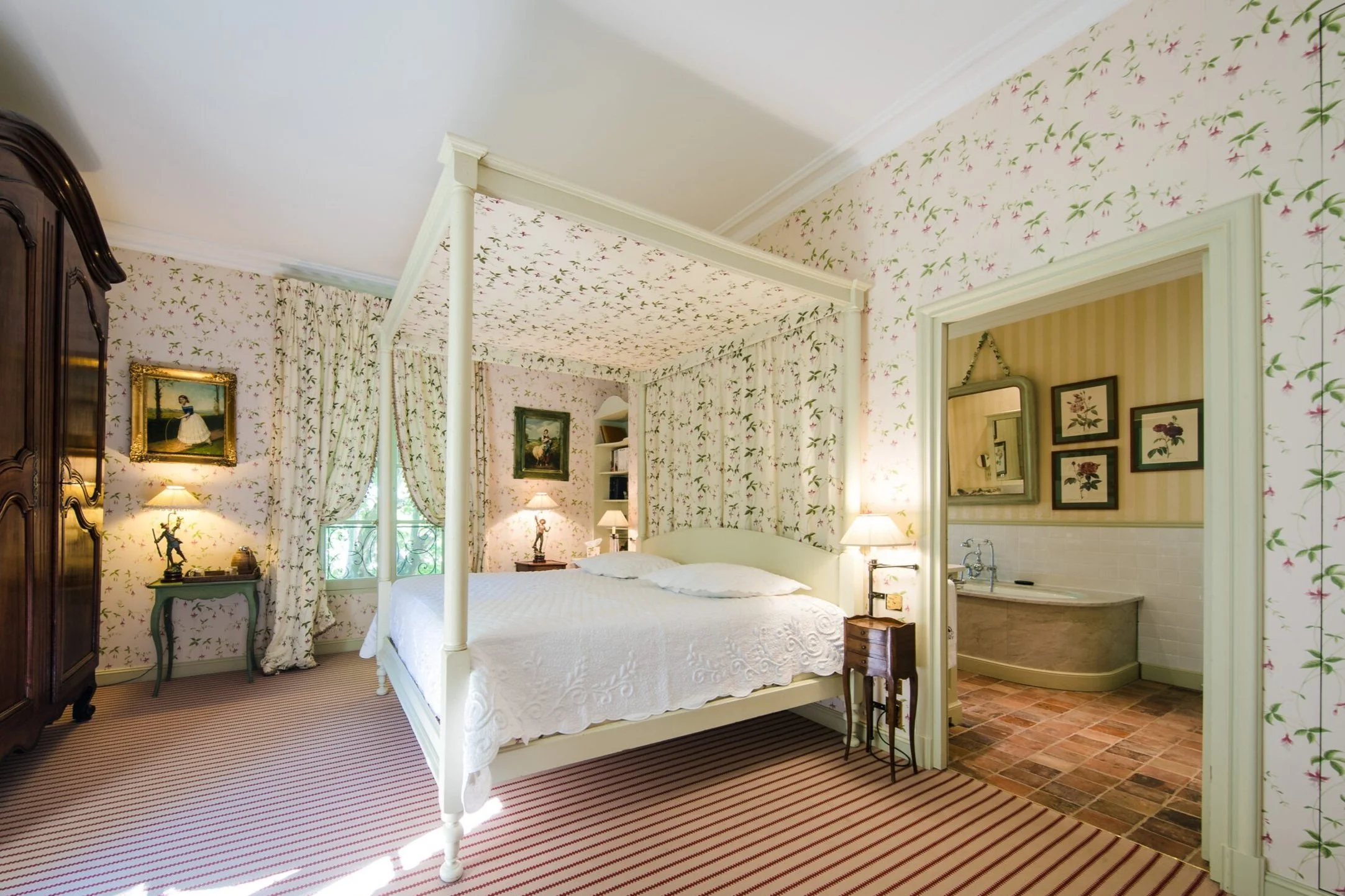A cozy bedroom with floral wallpaper, a white canopy bed, a wooden armoire, and a small bedside table with a lamp, leading into a bathroom with a bathtub and framed pictures on the wall.