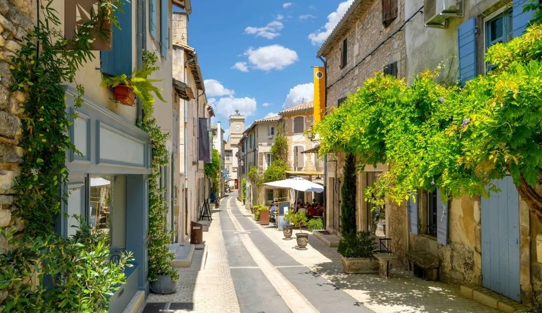 A narrow European street with stone buildings, outdoor seating with umbrellas, and lush green trees under a partly cloudy sky.
