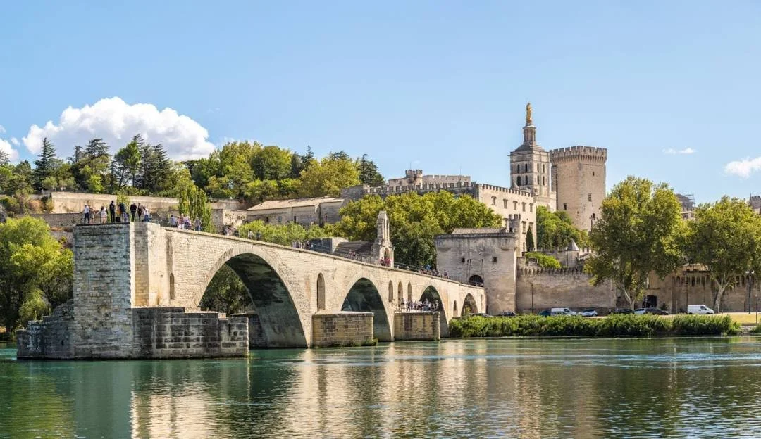 Historic stone bridge over a river with a castle and green trees in the background on a sunny day.