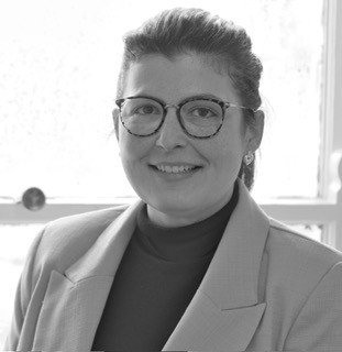 A woman with short hair wearing glasses, a blazer, and a turtleneck, smiling in front of a window.