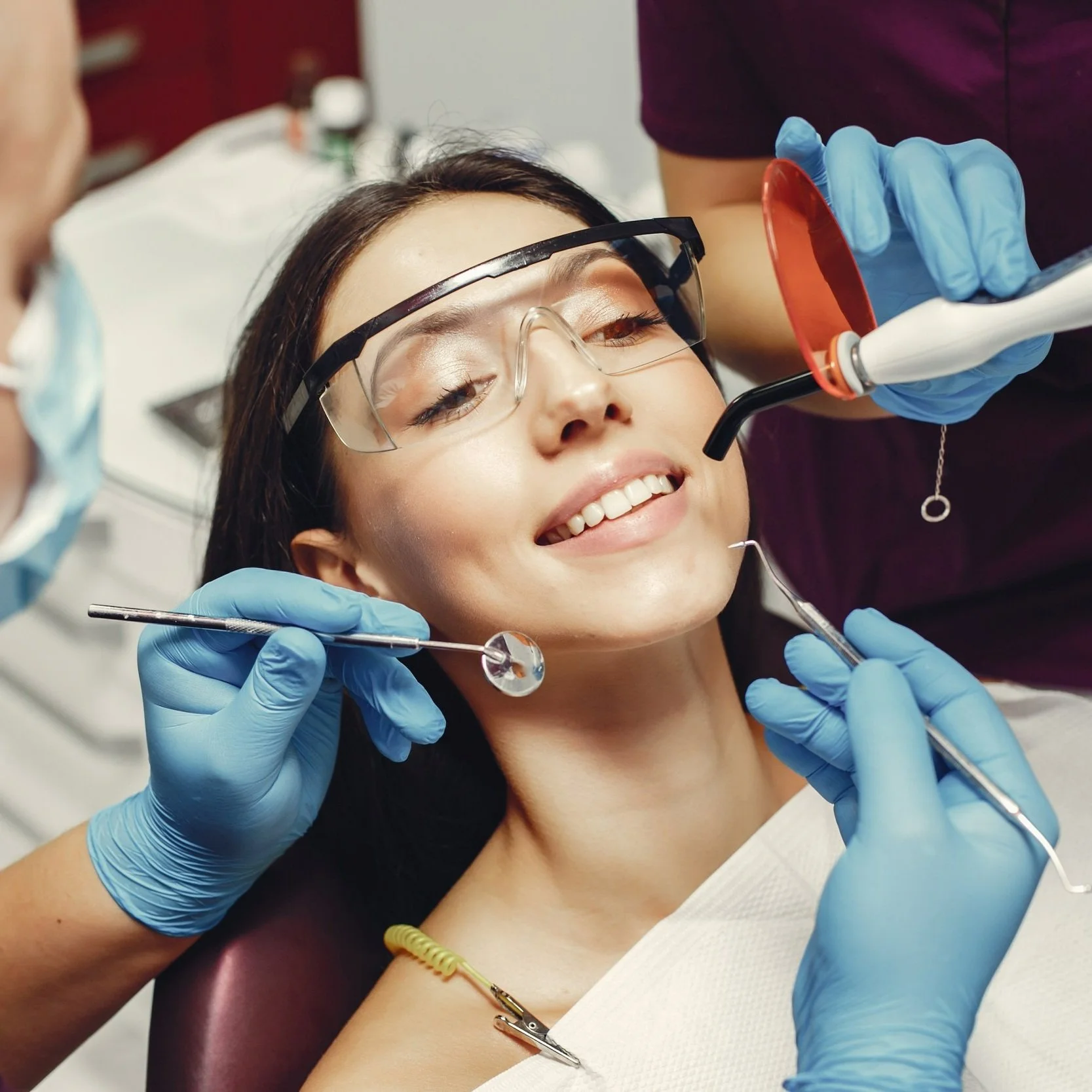 A woman at a dental clinic wearing protective glasses and smiling while receiving dental treatment. Dental tools and a dentist wearing blue gloves are visible.