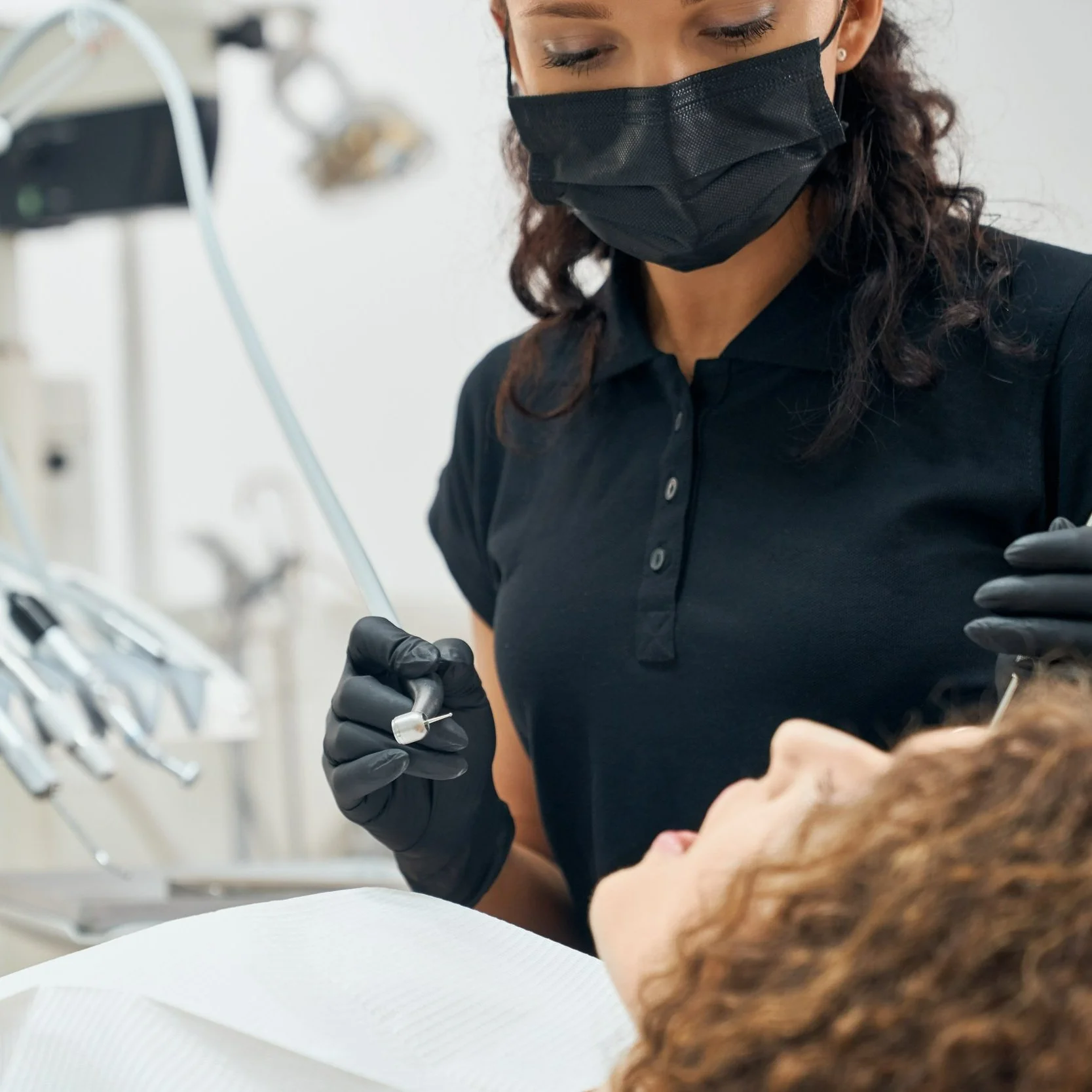 Dental professional wearing a black mask and gloves performing a dental procedure on a patient lying down in a dental chair.