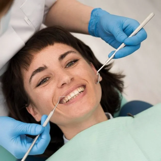 Young woman smiling during a dental check-up, lying in a dental chair with a dentist examining her mouth with dental mirrors.