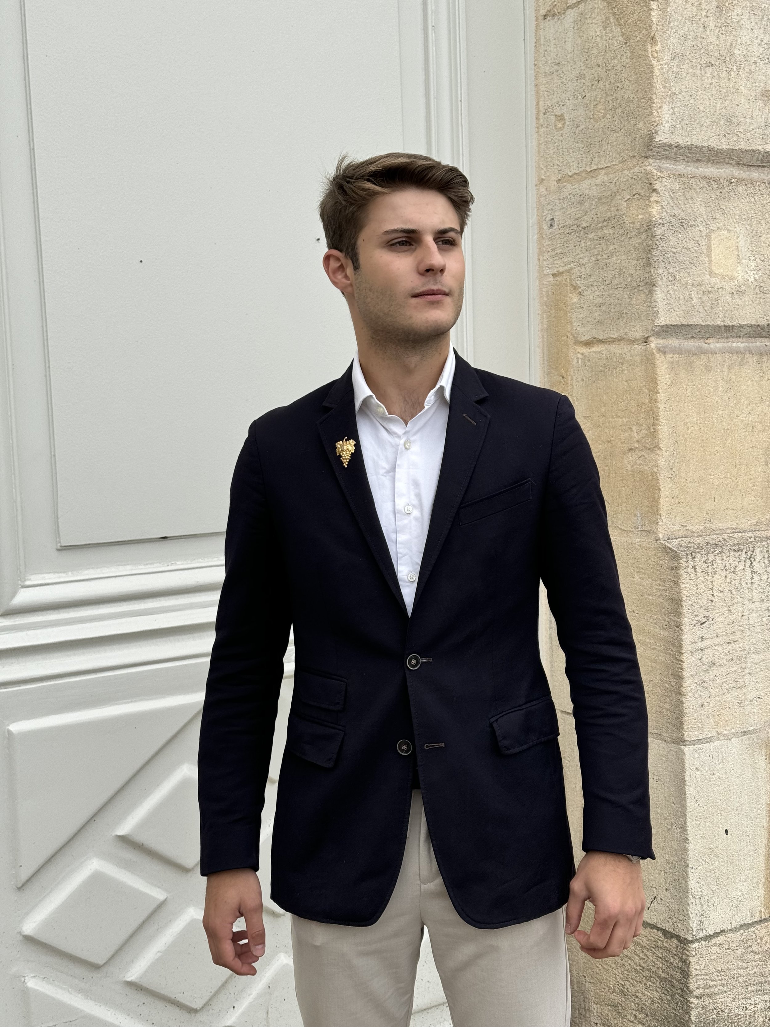Un jeune homme habillé en blazer noir, chemise blanche, et pantalon beige, se tenant devant un mur en pierre et une porte blanche.