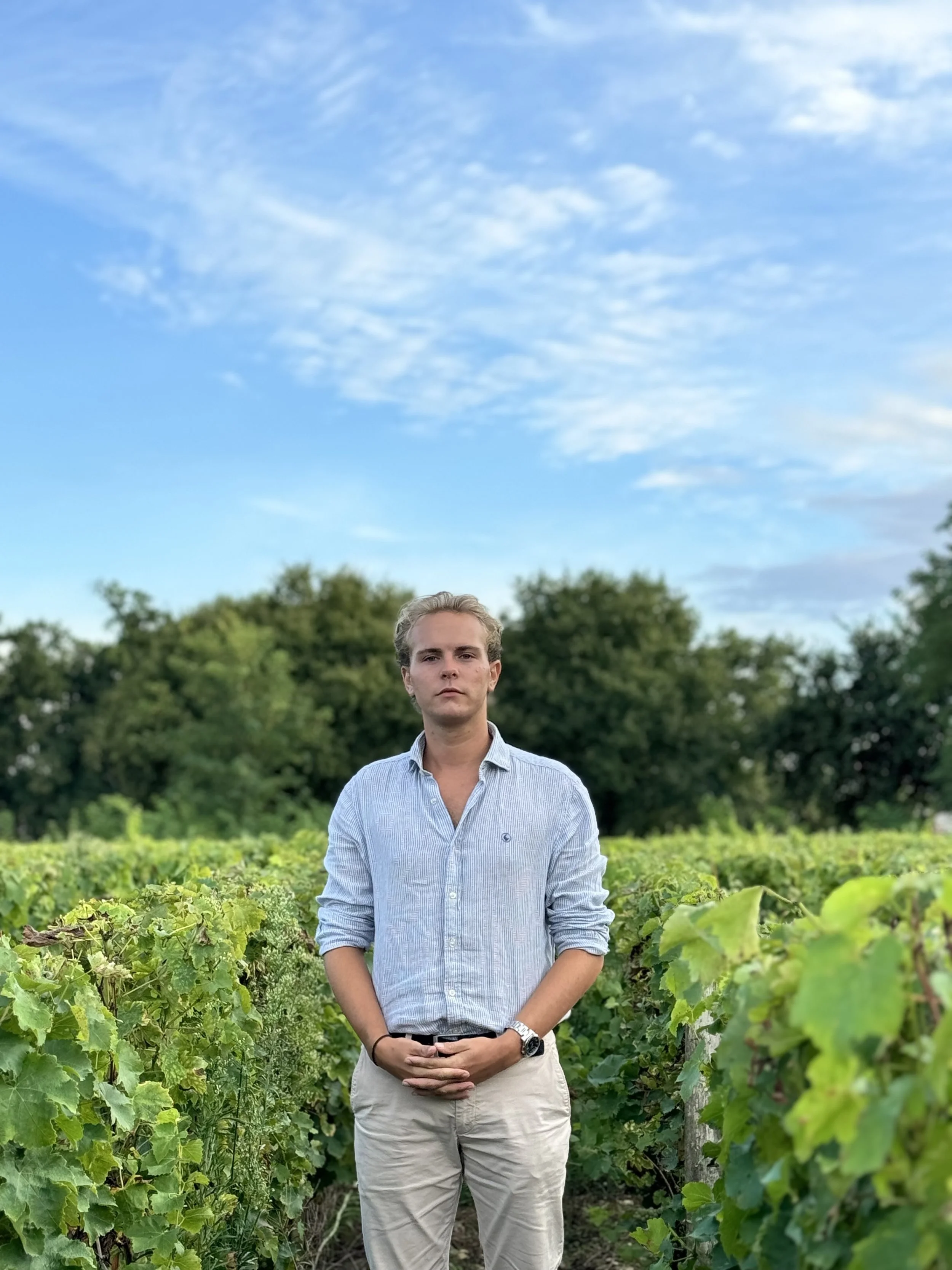Un jeune homme debout dans un vignoble avec un ciel bleu et quelques nuages.