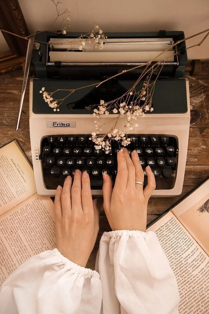 Hands typing on a vintage typewriter surrounded by open books and dried flowers
