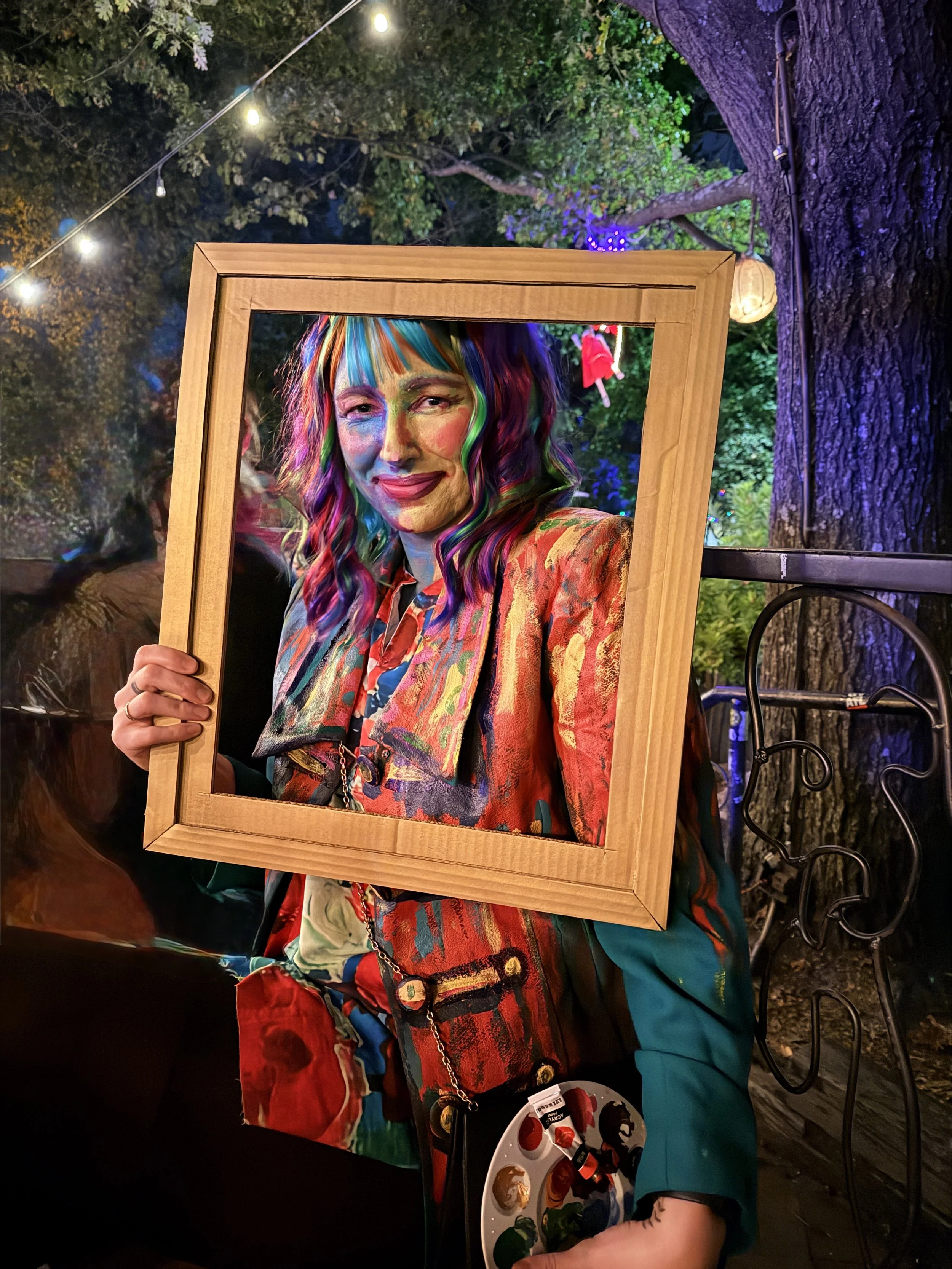 Person holding a wooden picture frame with a painted portrait of a woman with rainbow-colored hair, wearing a colorful jacket, at an outdoor nighttime event with string lights and trees in the background.