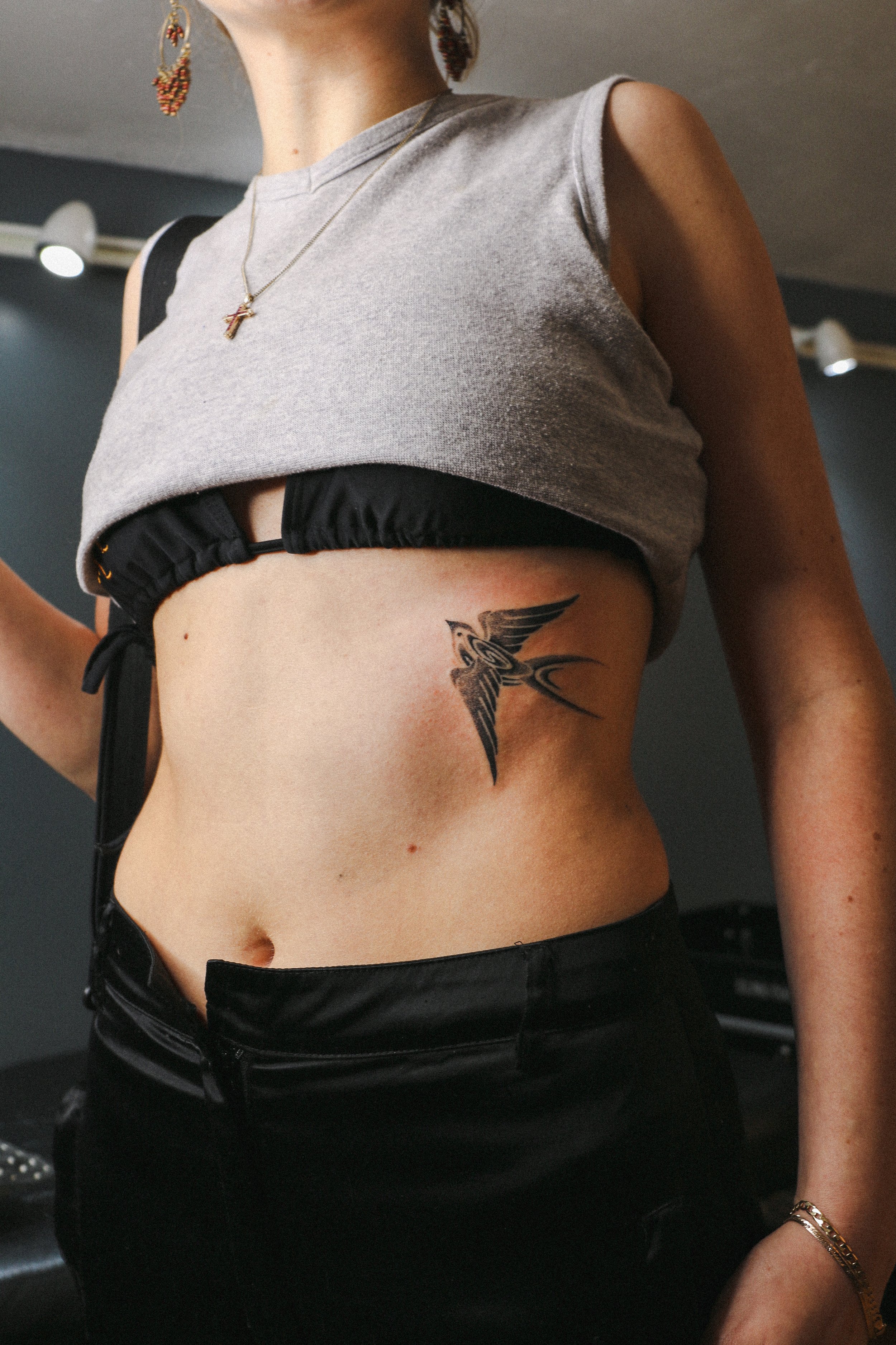 Person showing a bird tattoo on the right side of their torso, wearing a gray crop top and black pants.