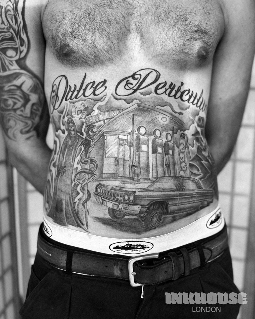 Chest and abdomen tattoos on a man, featuring "Dulce Periculum" text, a grim reaper, a car, and a building.